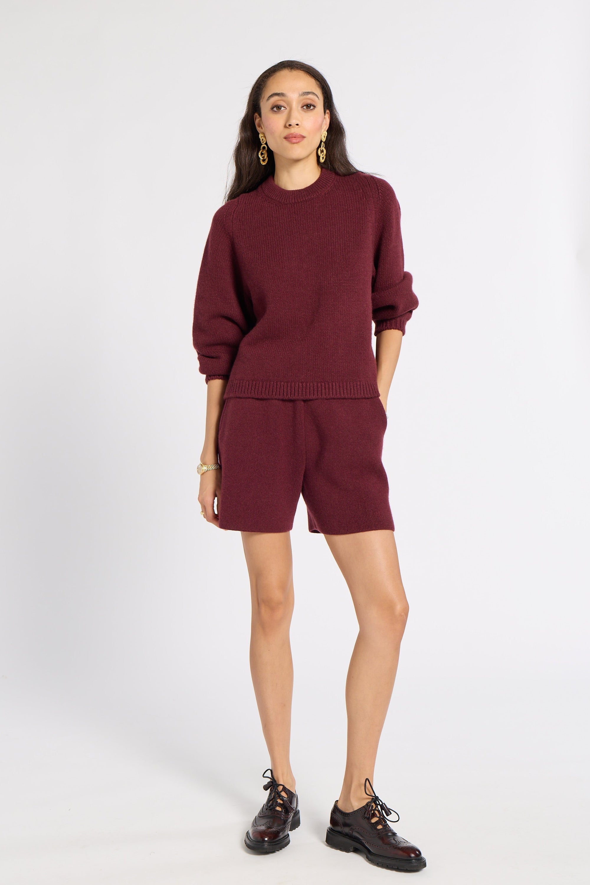 A woman modeling a matching burgundy knit sweater and shorts set, paired with dark brown leather lace-up shoes and gold hoop earrings, against a plain white studio background.