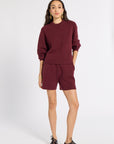 A woman modeling a matching burgundy knit sweater and shorts set, paired with dark brown leather lace-up shoes and gold hoop earrings, against a plain white studio background.