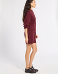A woman stands in profile wearing a matching burgundy knit sweater and shorts set, paired with dark brown leather oxford shoes, against a plain light gray studio background.
