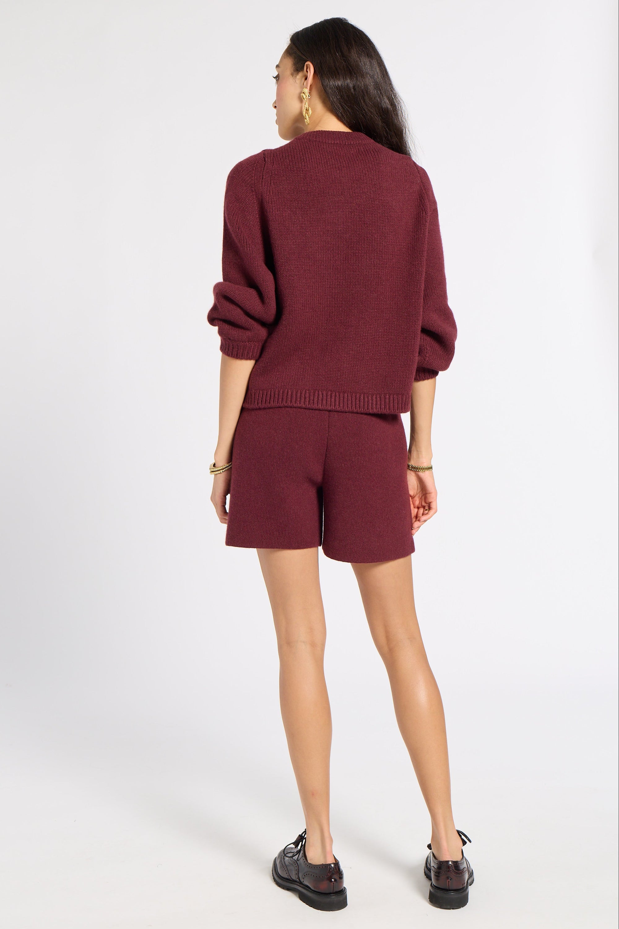 A woman shown from the back wearing a matching burgundy knit sweater and shorts set, paired with black shoes, against a plain light gray studio background.
