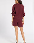 A woman shown from the back wearing a matching burgundy knit sweater and shorts set, paired with black shoes, against a plain light gray studio background.