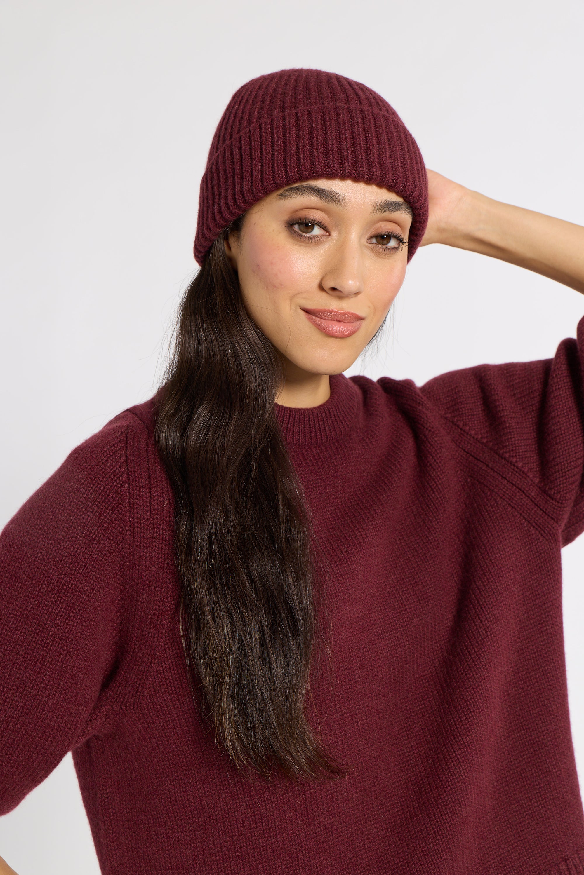A woman wearing a ribbed cashmere beanie and a matching ribbed knit sweater, posed against a plain light gray studio background.