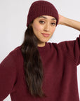 A woman wearing a ribbed cashmere beanie and a matching ribbed knit sweater, posed against a plain light gray studio background.