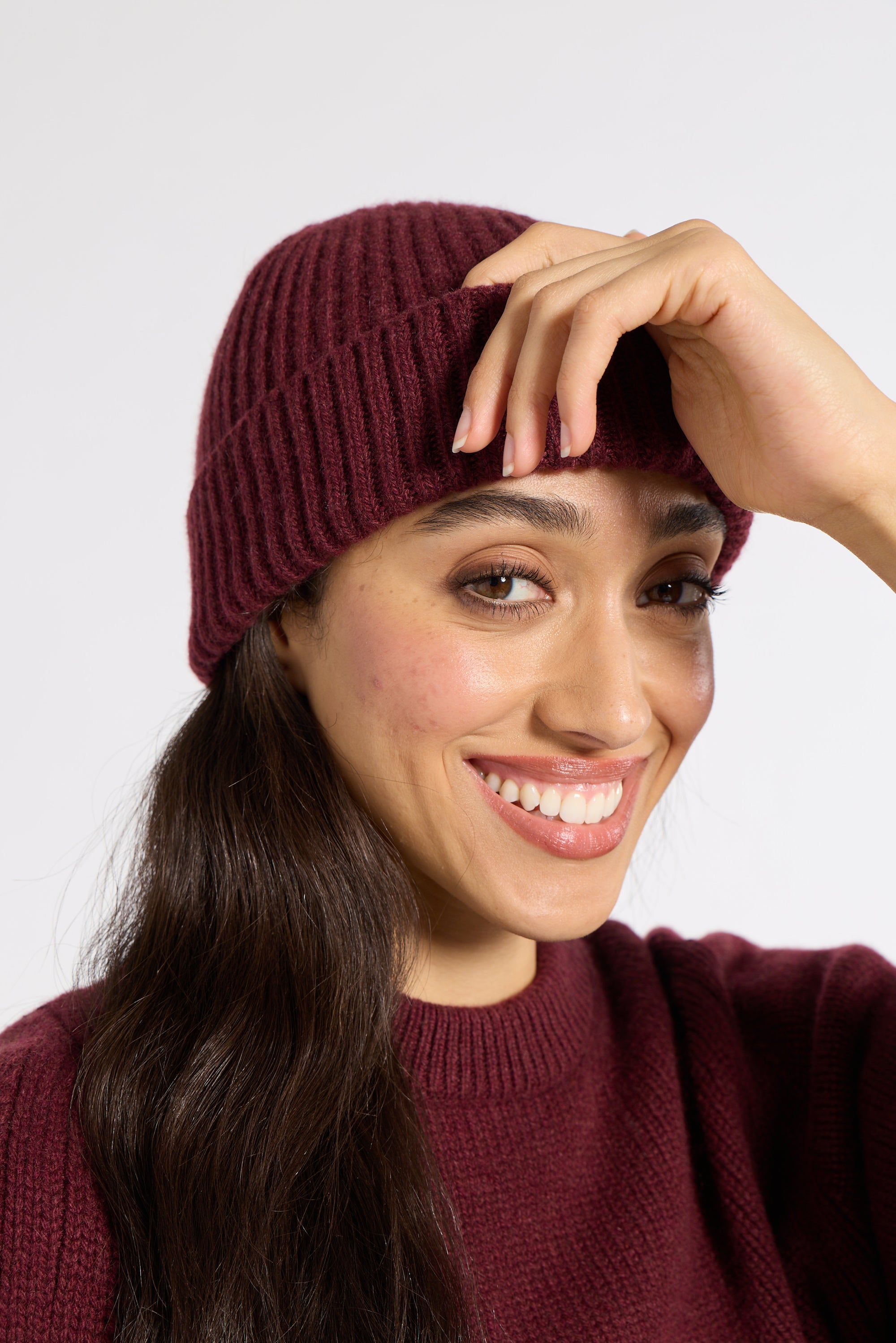 A smiling woman with long dark hair wearing a ribbed knit beanie and matching knit sweater against a plain light gray background.