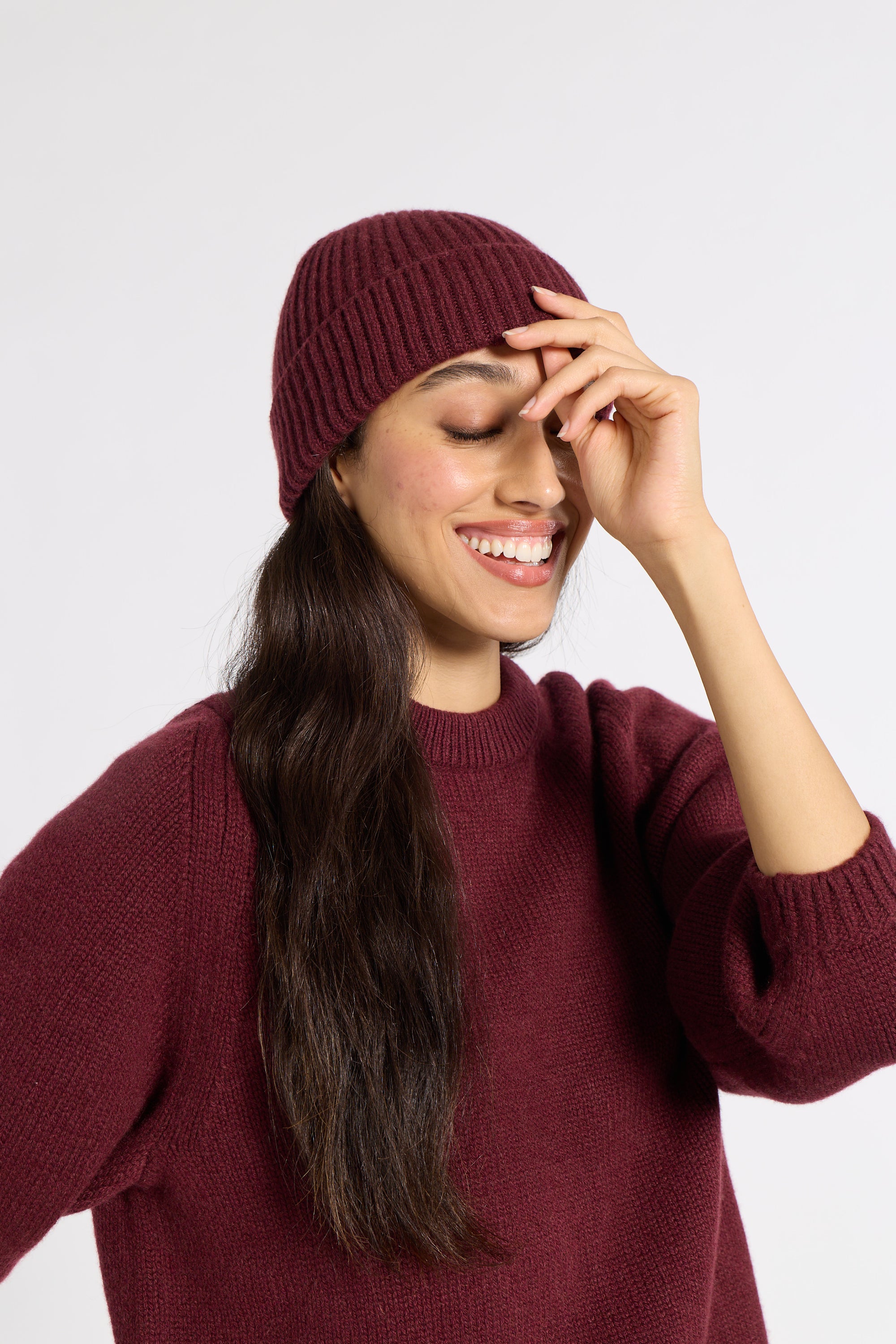A woman wearing a ribbed burgundy knit beanie and a matching burgundy knit sweater, smiling against a light gray studio background.