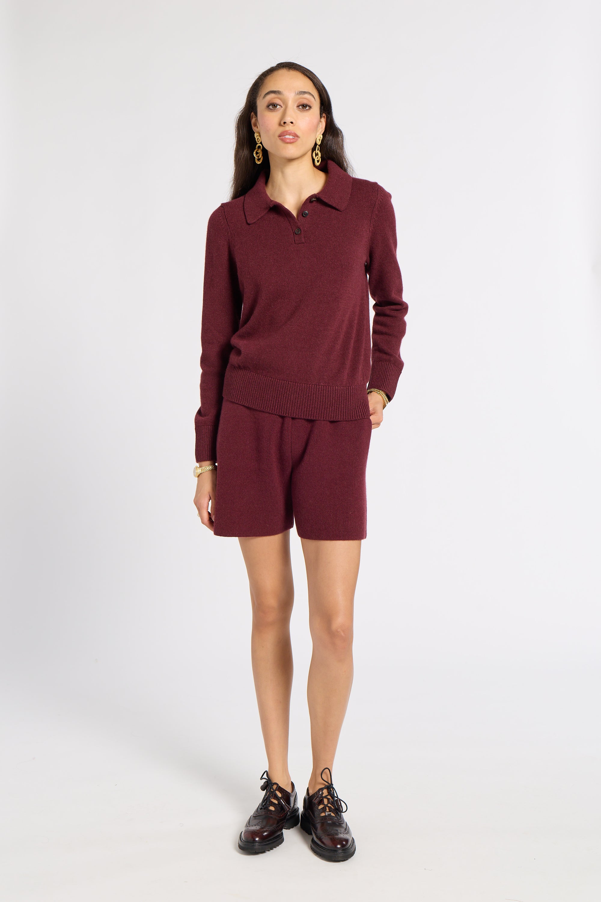 A woman modeling a matching burgundy knit set consisting of a long-sleeve polo shirt and shorts, paired with black lace-up shoes against a plain white studio background.