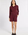 A woman modeling a matching burgundy knit set consisting of a long-sleeve polo shirt and shorts, paired with black lace-up shoes against a plain white studio background.