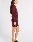 A woman in profile wearing a matching burgundy knit long-sleeve top and shorts set, paired with dark brown leather oxford shoes, against a plain light gray studio background.