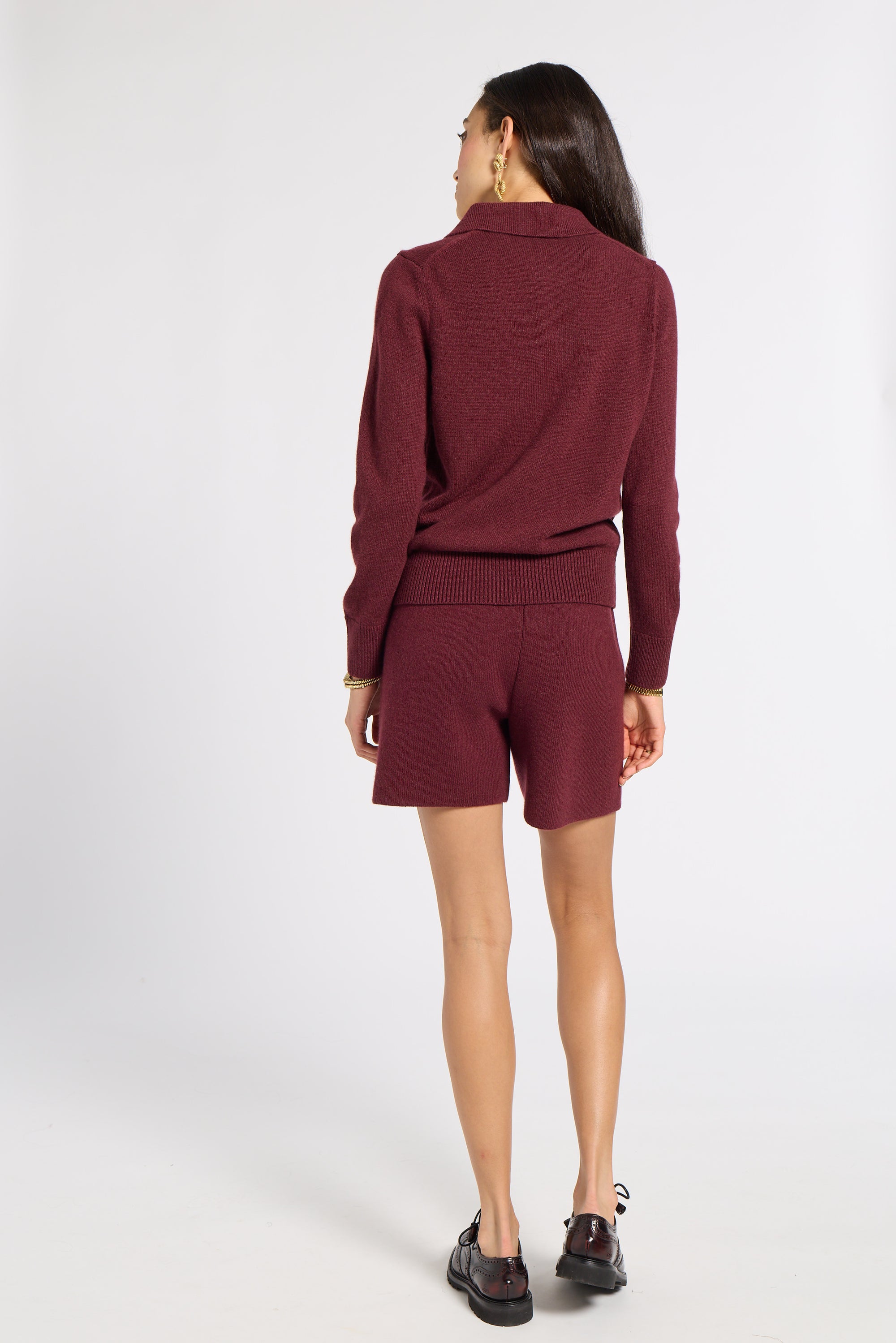 A woman shown from the back wearing a matching burgundy knit set consisting of a long-sleeve polo-style top and shorts, paired with dark loafers, against a light gray studio background.