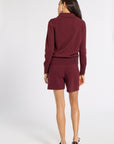 A woman shown from the back wearing a matching burgundy knit set consisting of a long-sleeve polo-style top and shorts, paired with dark loafers, against a light gray studio background.