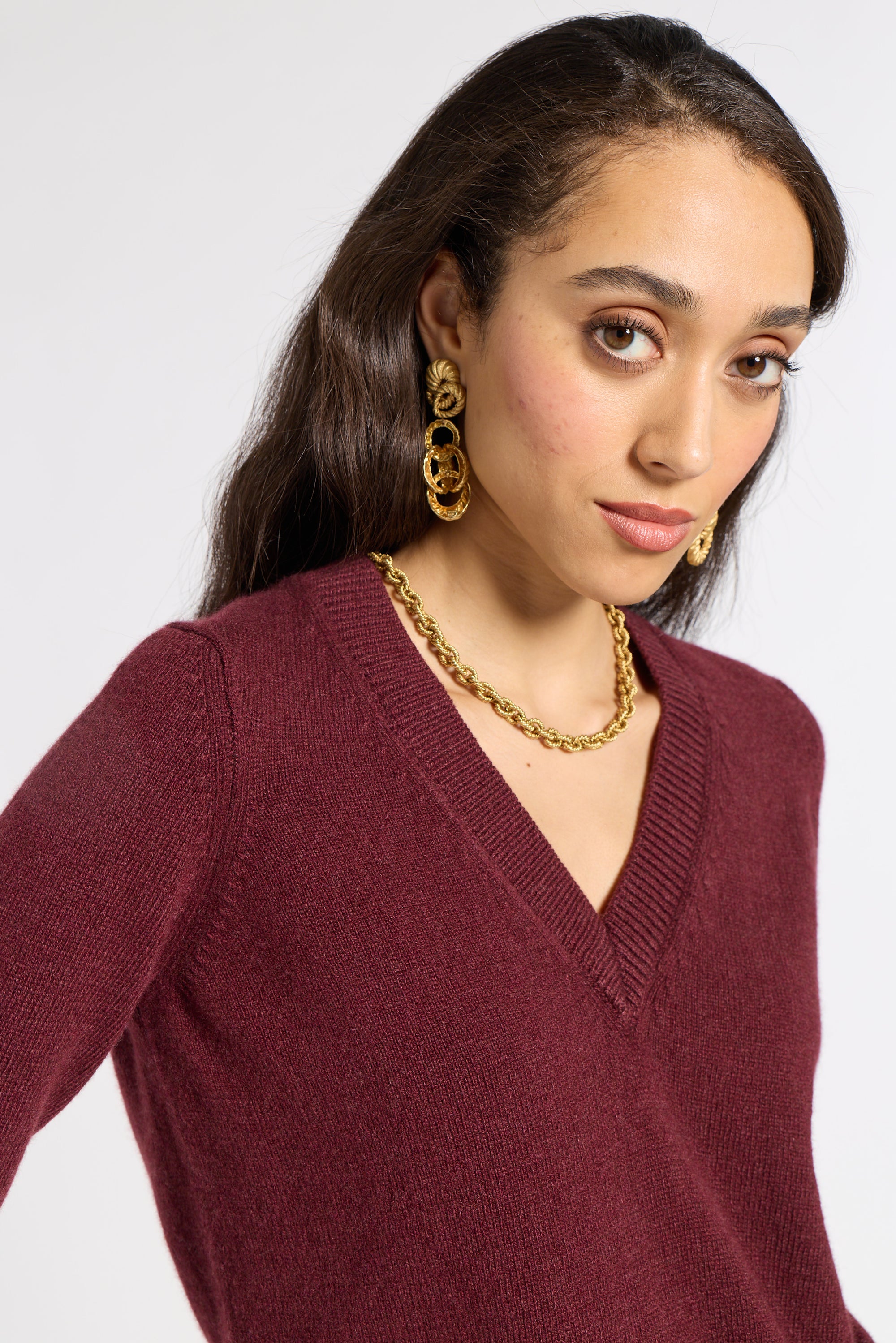 woman modeling a burgundy v neck sweater with gold jewelry on a white backgroun