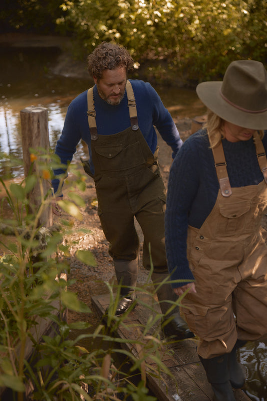 Couple wearing outdoor gear, including the Men's Redford Crew sweater in navy, walking through a forest near a stream during a nature activity.