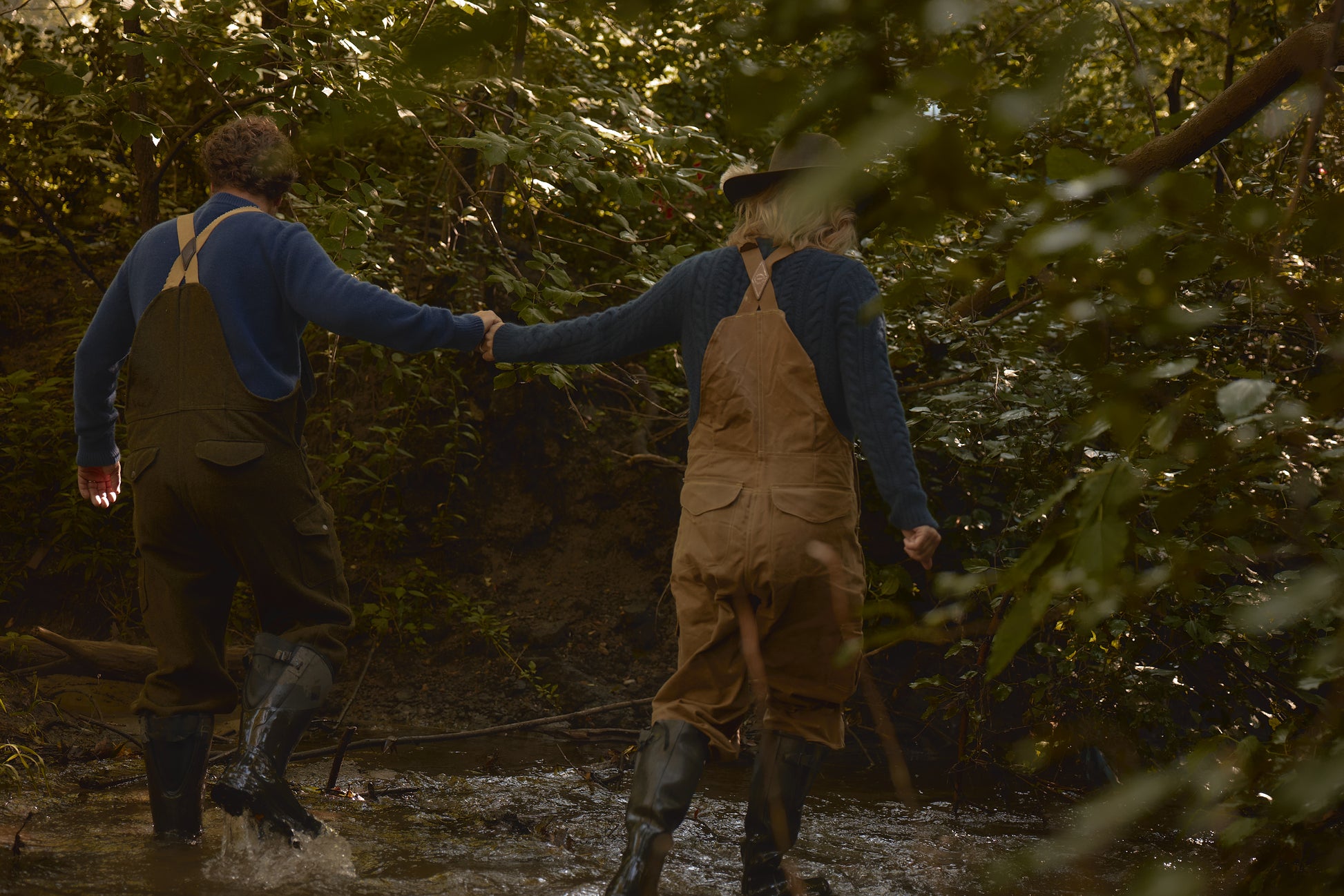 Two people wearing outdoor gear, including the Men's Redford Crew in navy, walking hand in hand through a shallow stream in a forest setting.