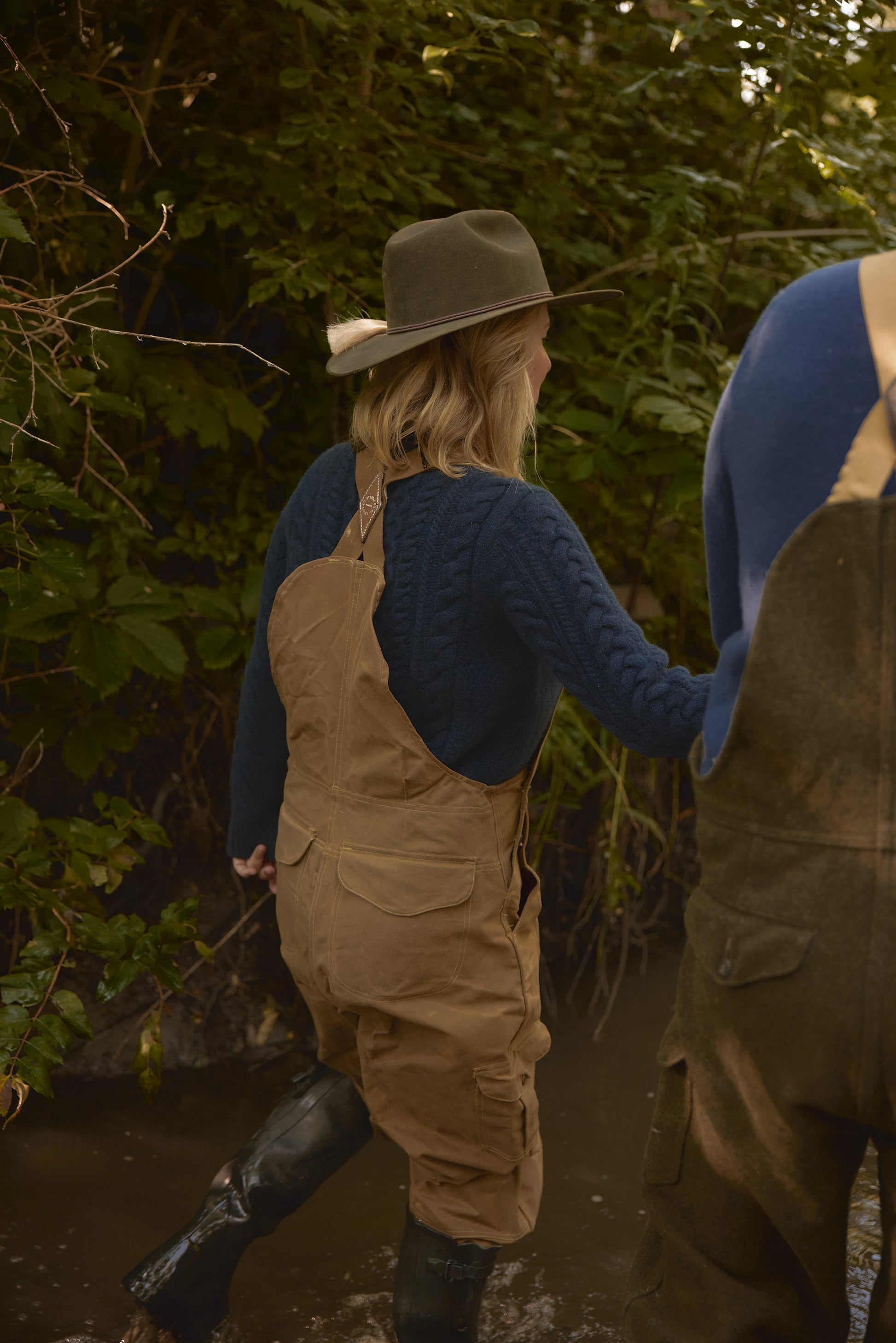 Eleanor Leftwich wading through a stream in the Navy Fisherman Crewneck, tan waterproof overalls, tall black boots, and a faded green hat.