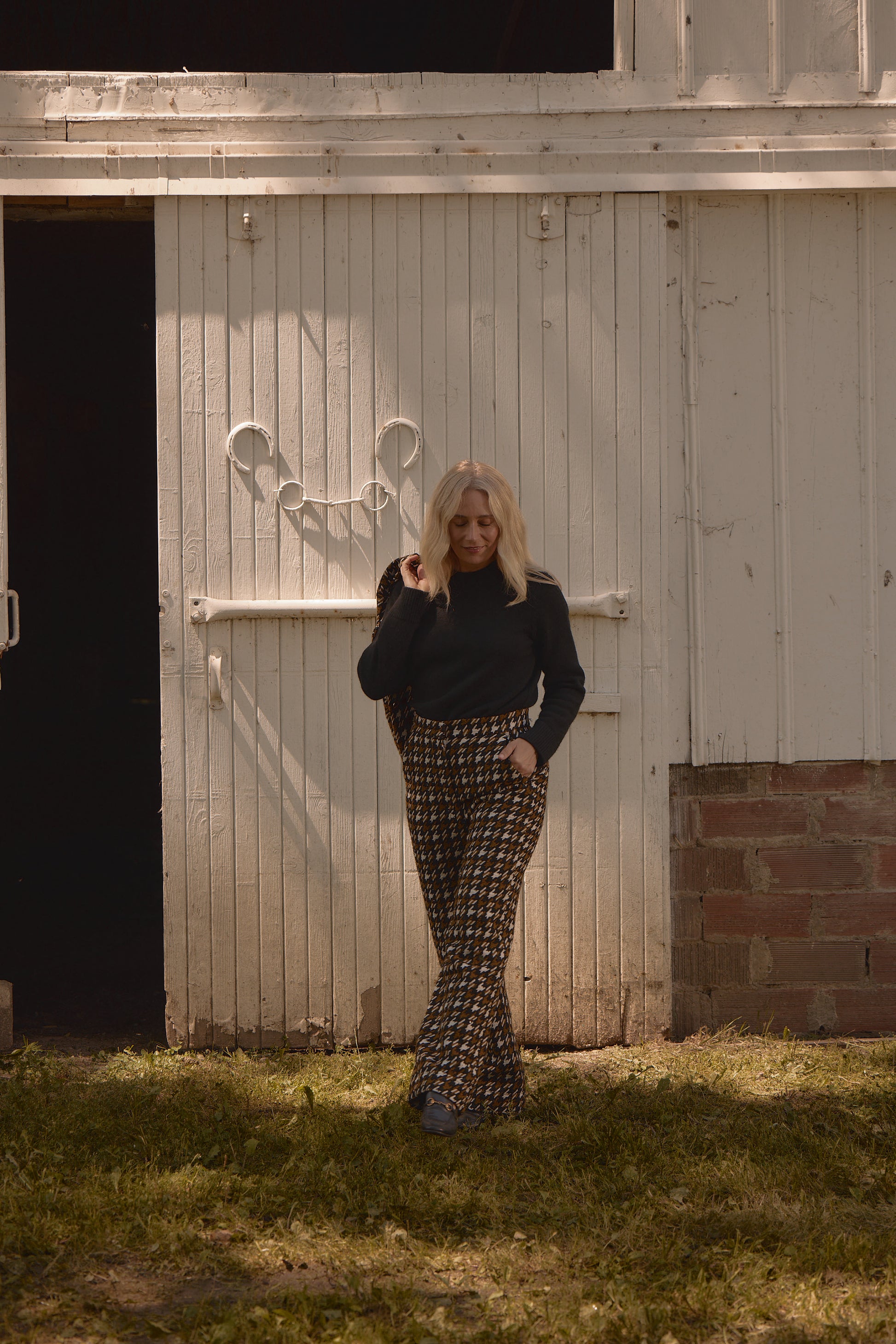 Model wearing the Classic Cashmere Crew in black, paired with brown houndstooth trousers, standing outdoors against a rustic wooden barn door.