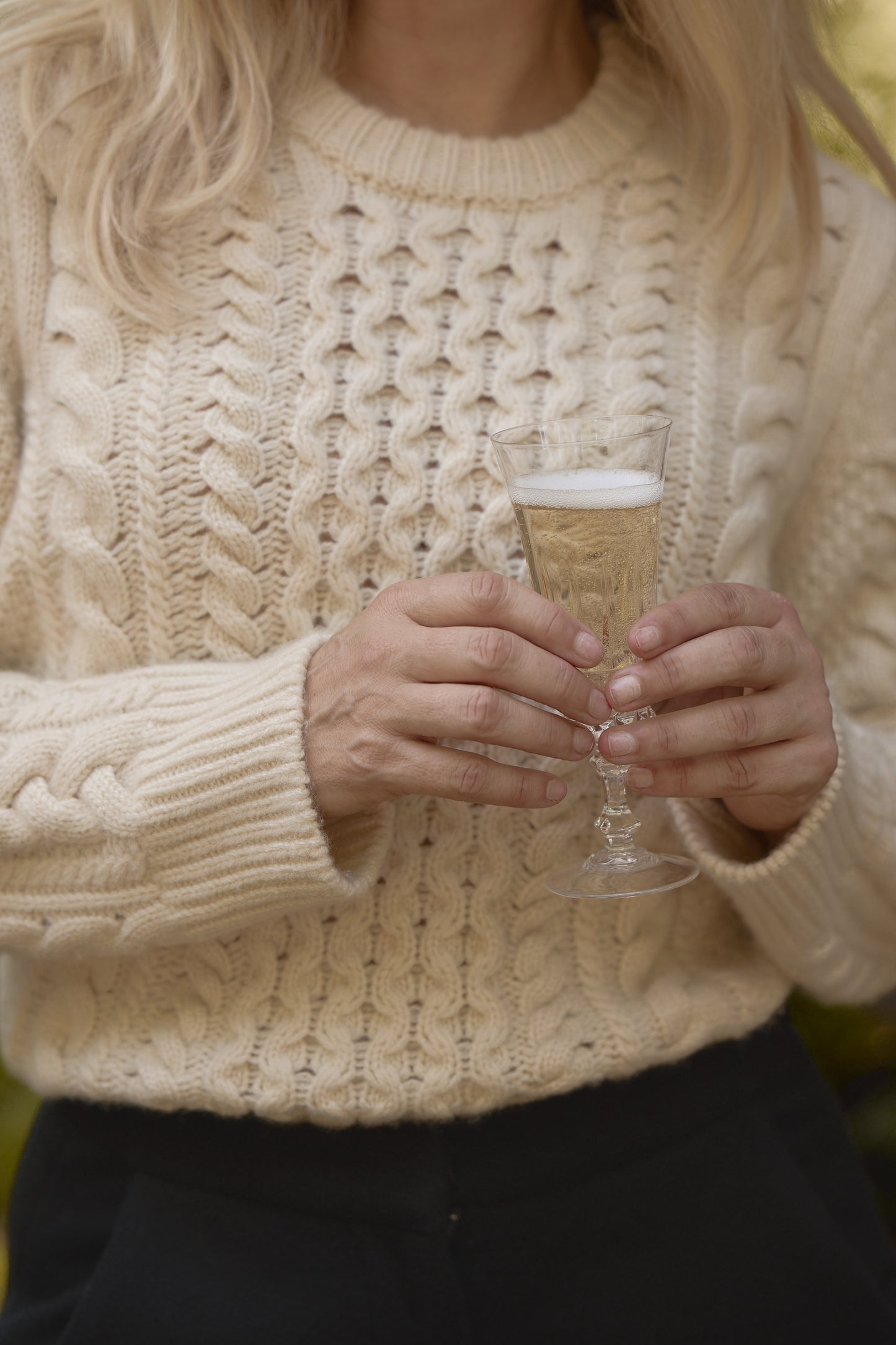 Close-up shot of model holding a glass of champagne in the cream chunky cable knit fisherman crewneck, paired with black slacks.