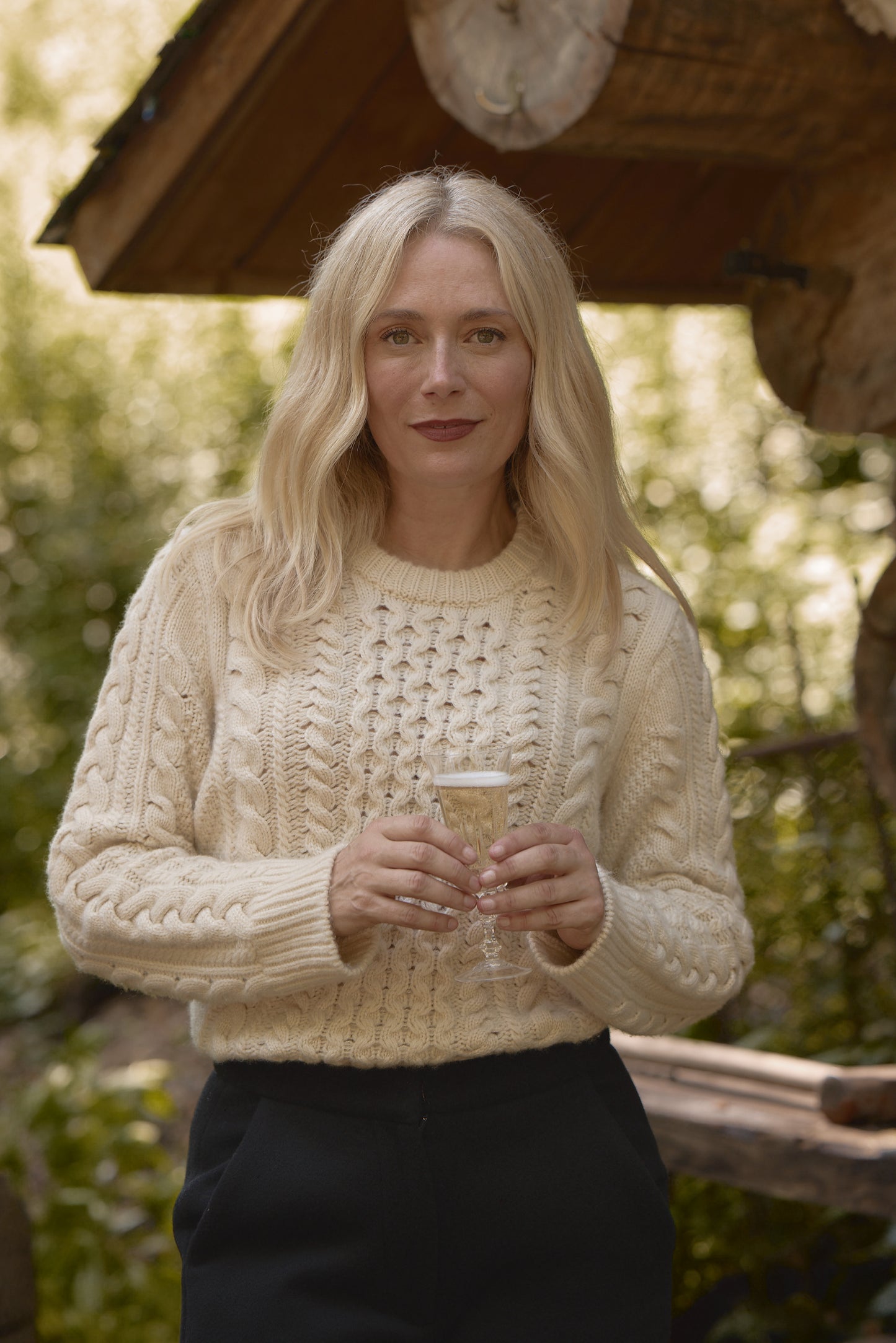 Blonde woman wearing the chunky Aran cable knit fisherman crewneck in cream, holding a glass of champagne in an outdoor, rustic setting.