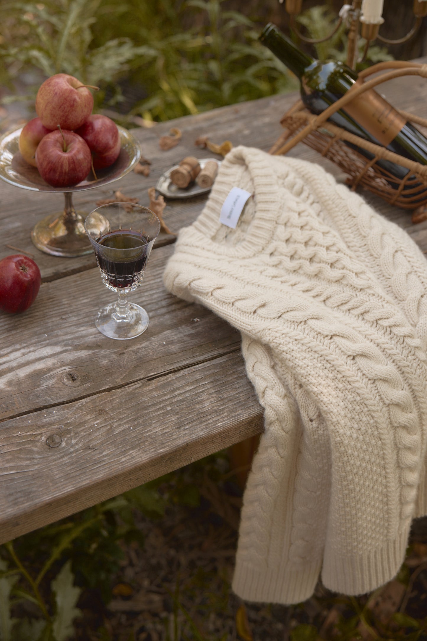 Chunky Aran cable knit fisherman crewneck in cream on a rustic table with apples and a glass of wine, creating a cozy autumn scene