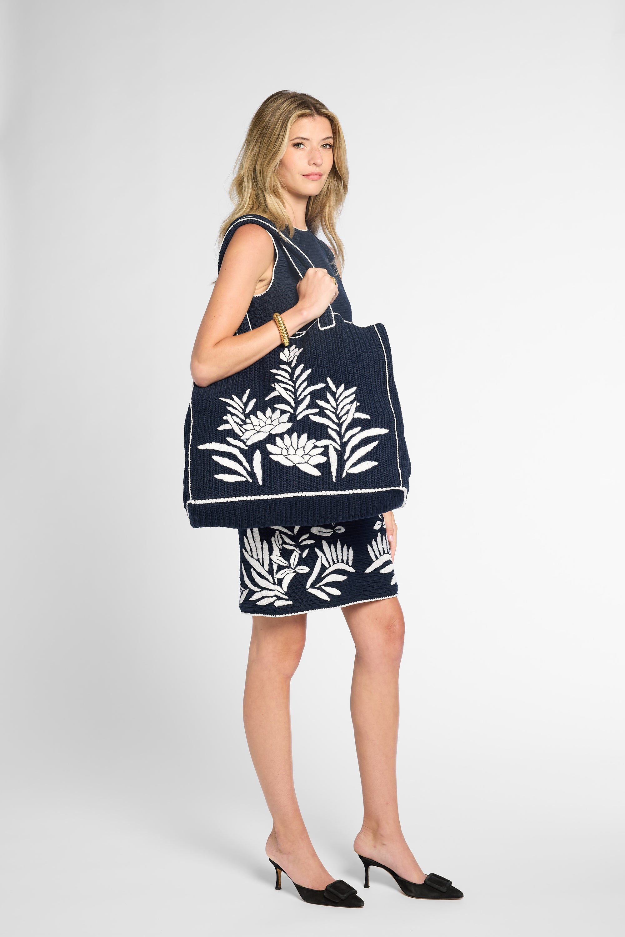 A woman wearing a navy black crochet dress and holding a matching black crochet  tote bag with white floral embroidery, against a light gray studio background. Styled with black mule pumps.