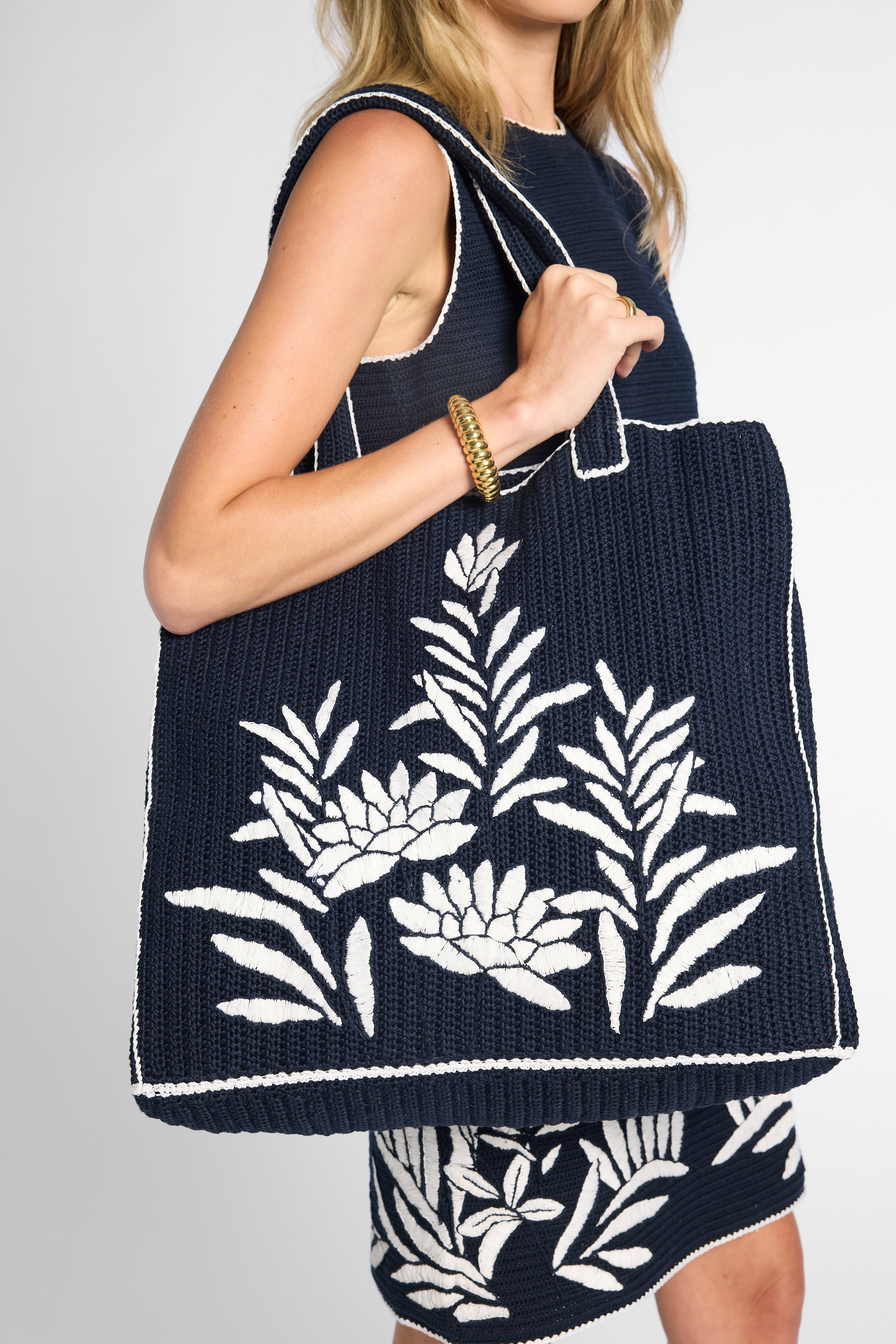 A woman wearing a navy black crochet dress and holding a matching black crochet  tote bag with white floral embroidery, against a light gray studio background.
