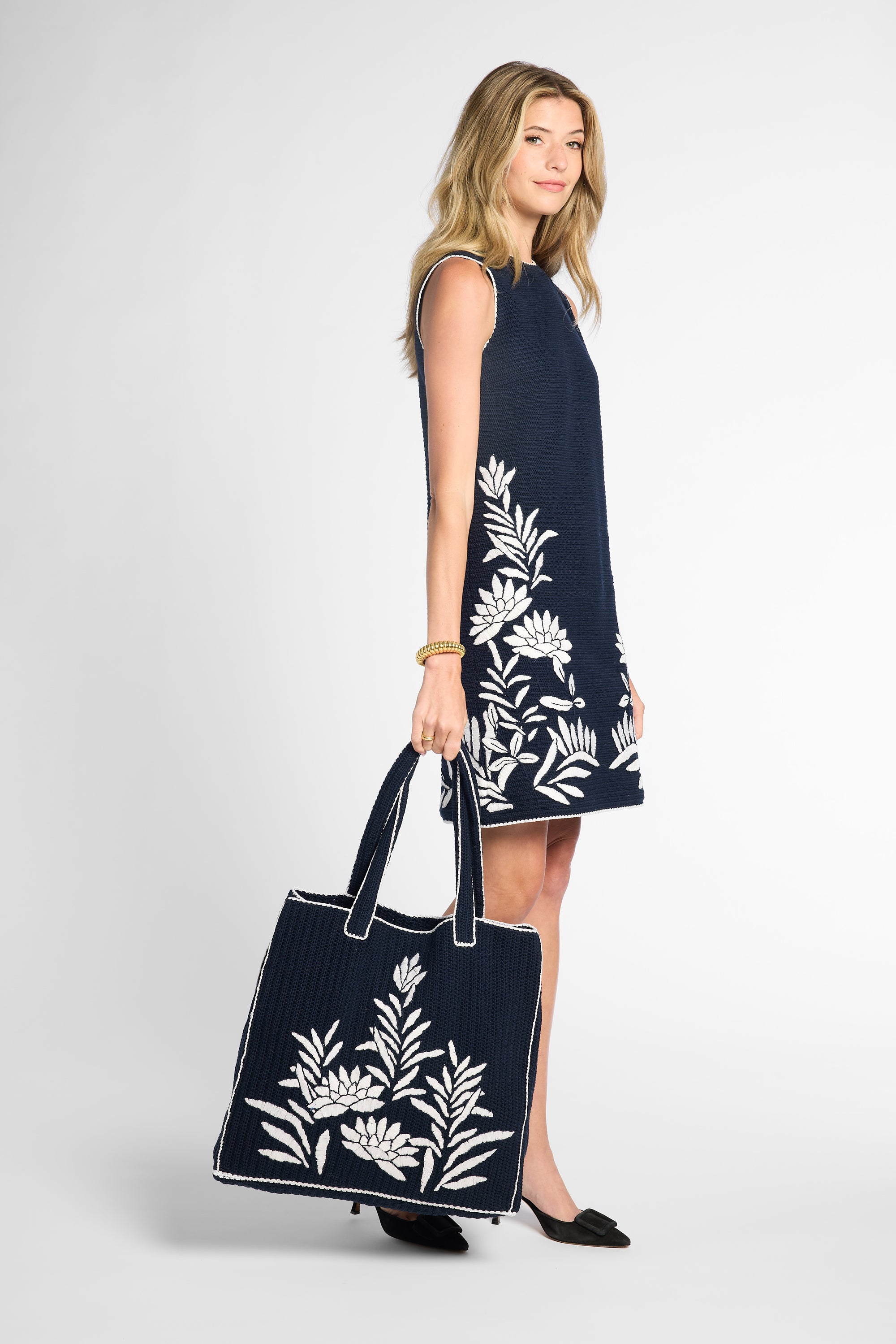 A woman stands in profile wearing a sleeveless black dress with large white floral embroidery and holding a matching black tote bag with white floral embroidery, against a light gray studio background.