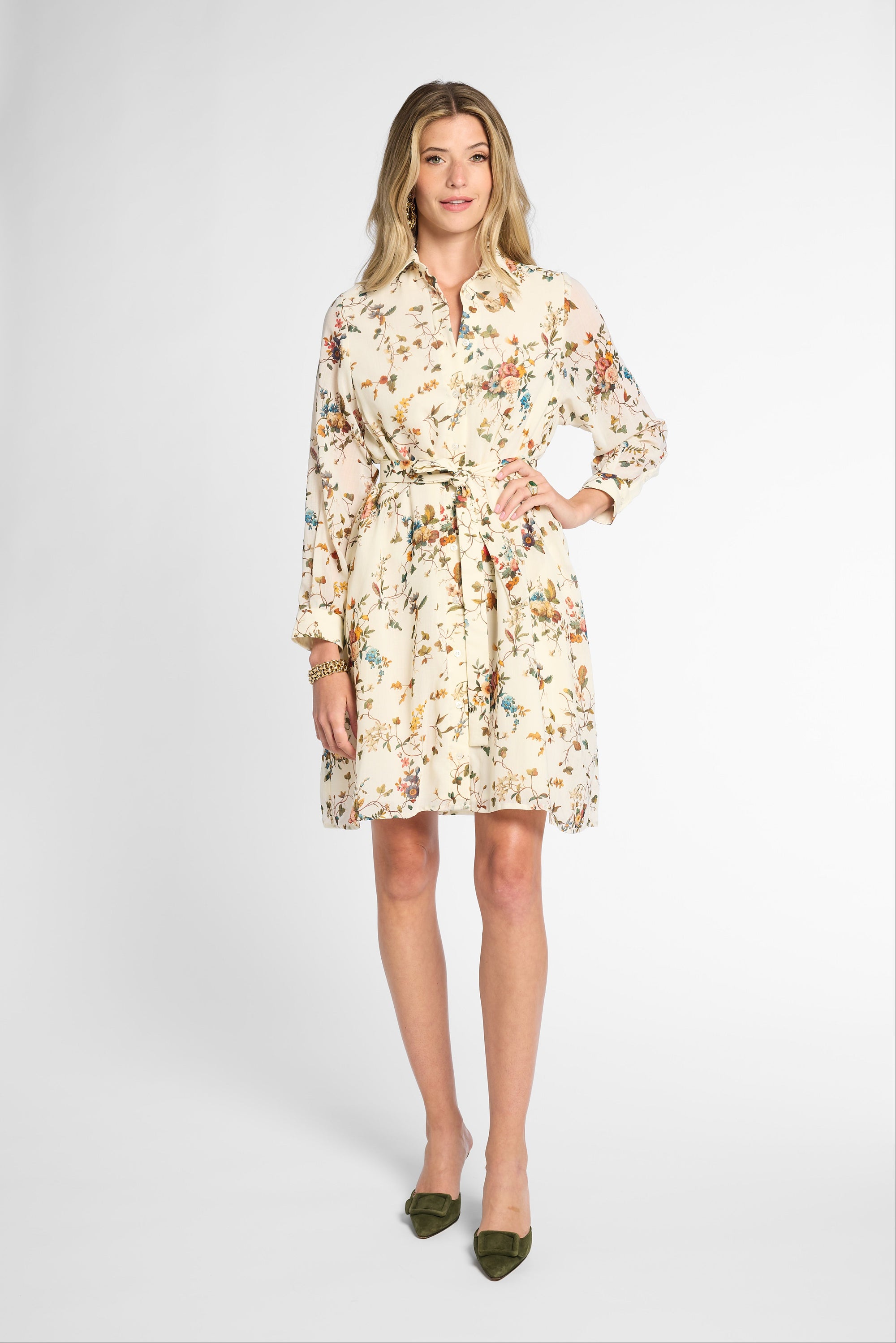 A woman modeling a long-sleeve cream-colored shirt dress with a multicolor floral print and a matching tie belt, paired with olive green pointed-toe flats, against a light gray studio background.