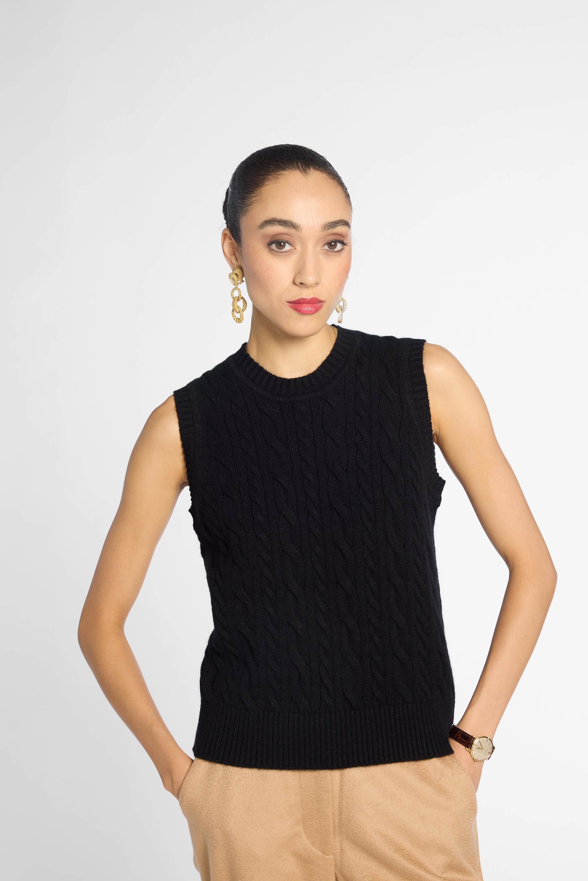 A woman modeling a sleeveless black cable-knit sweater vest with a mock neck, paired with beige trousers, against a light gray studio background.