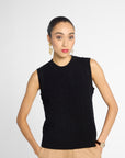 A woman modeling a sleeveless black cable-knit sweater vest with a mock neck, paired with beige trousers, against a light gray studio background.