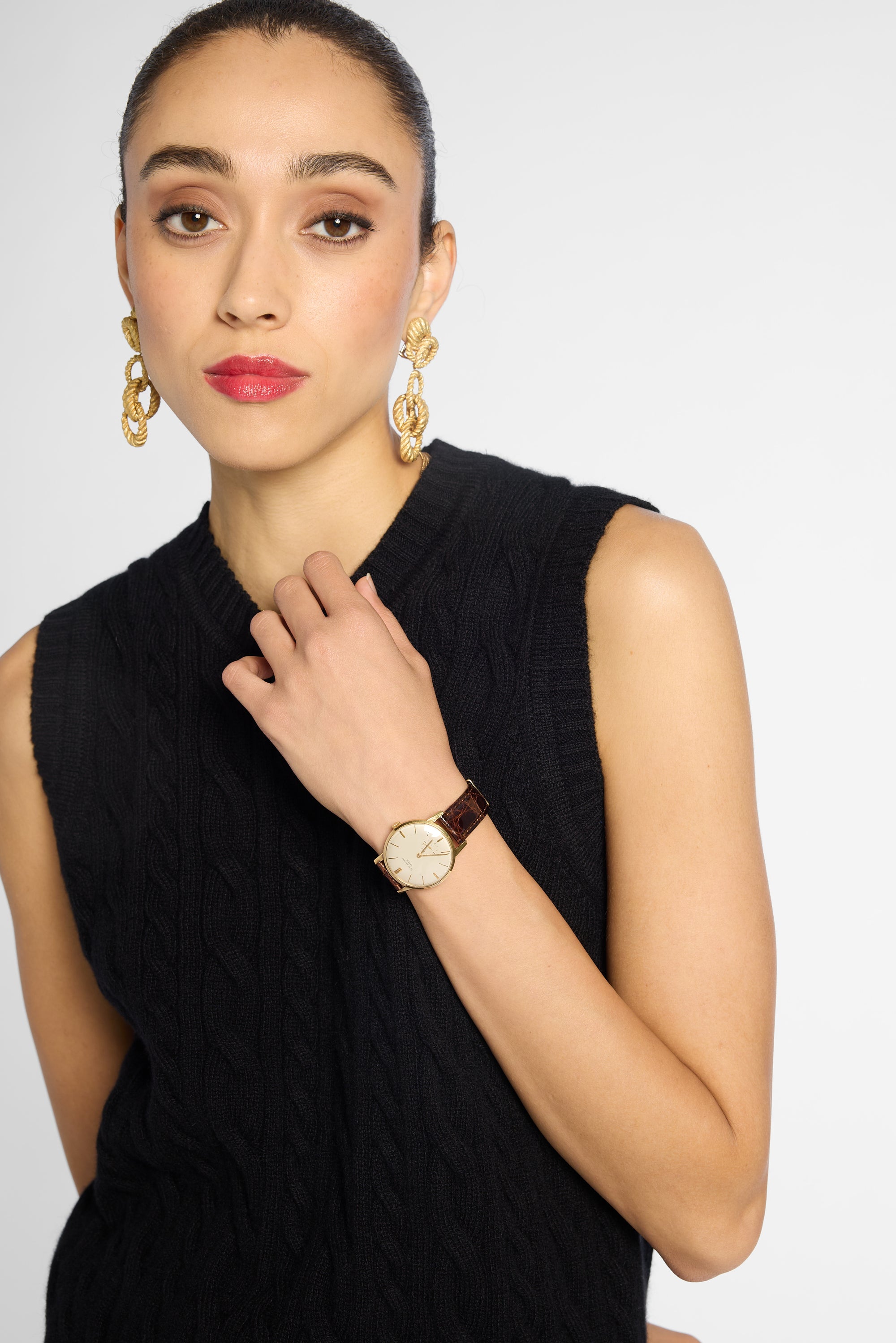 A woman wearing a black sleeveless cable-knit top and a gold-tone wristwatch with a cream dial and brown leather strap, posed against a light gray studio background.