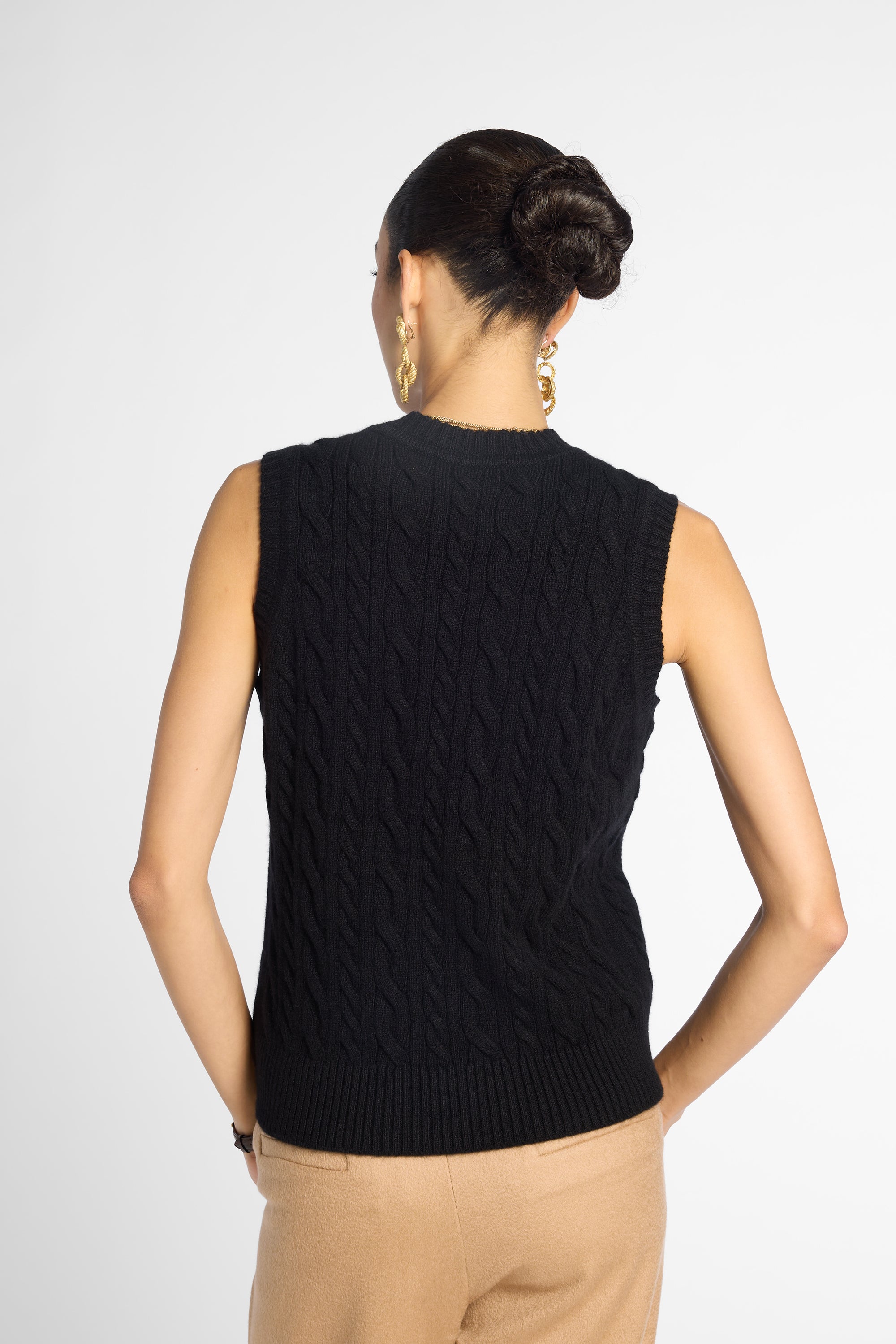 A woman shown from the back wearing a sleeveless black cable-knit sweater vest and beige pants against a light gray studio background.