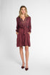 A woman modeling a long-sleeve, wrap-style mini dress with vertical pleats in a deep burgundy color, paired with matching pointed-toe heels, against a light gray studio background.
