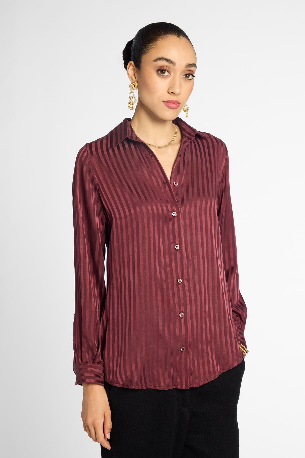 A woman wearing a long-sleeve button-up blouse with vertical stripes in a deep burgundy and maroon color, paired with black pants, against a plain light gray background.

