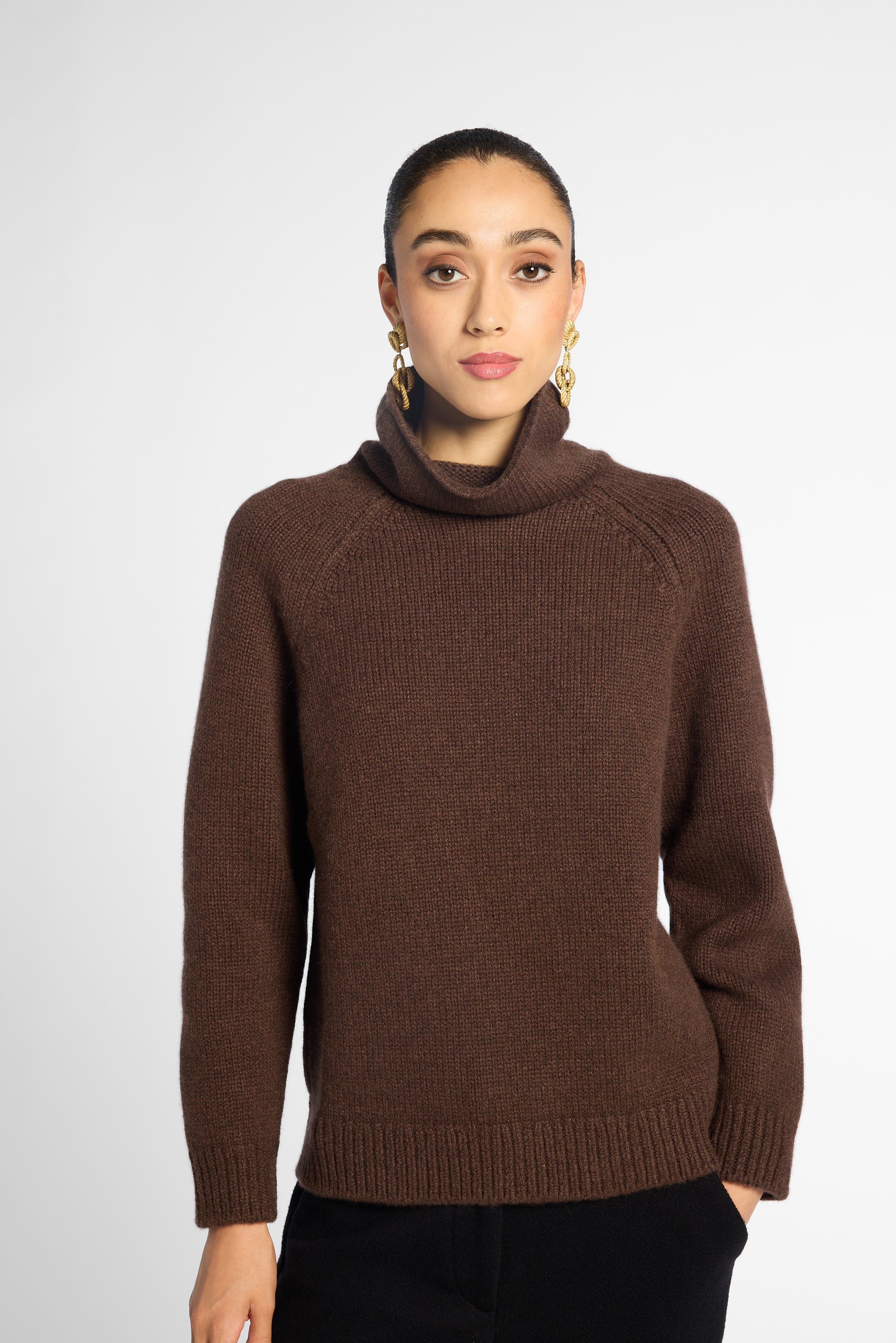 Front view: woman wearing a turtleneck sweater in Chocolate brown with black pants in front of a white background 