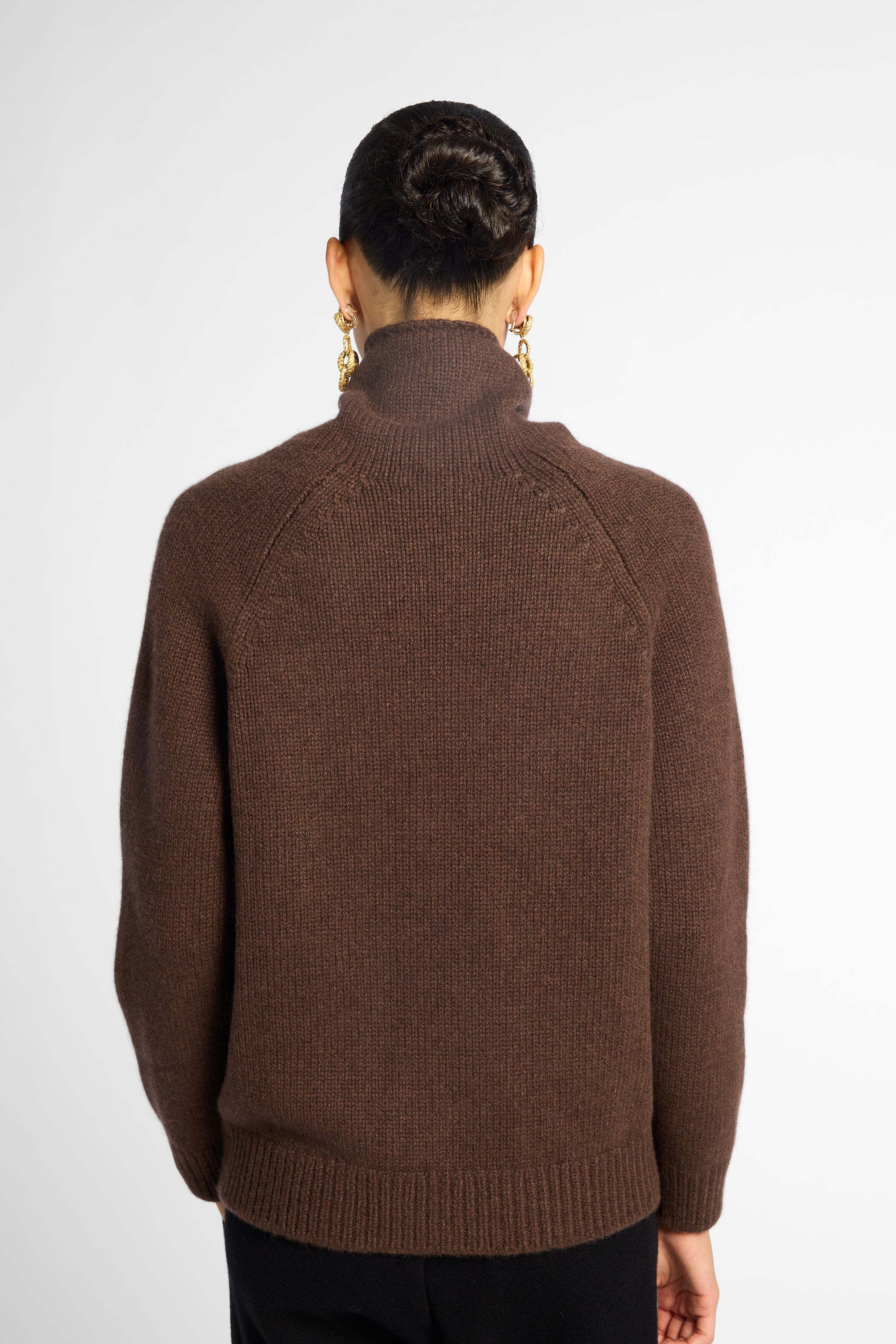 Back view: woman wearing a turtleneck sweater in Chocolate brown with black pants in front of a white background 