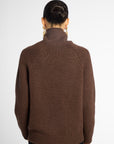Back view: woman wearing a turtleneck sweater in Chocolate brown with black pants in front of a white background 