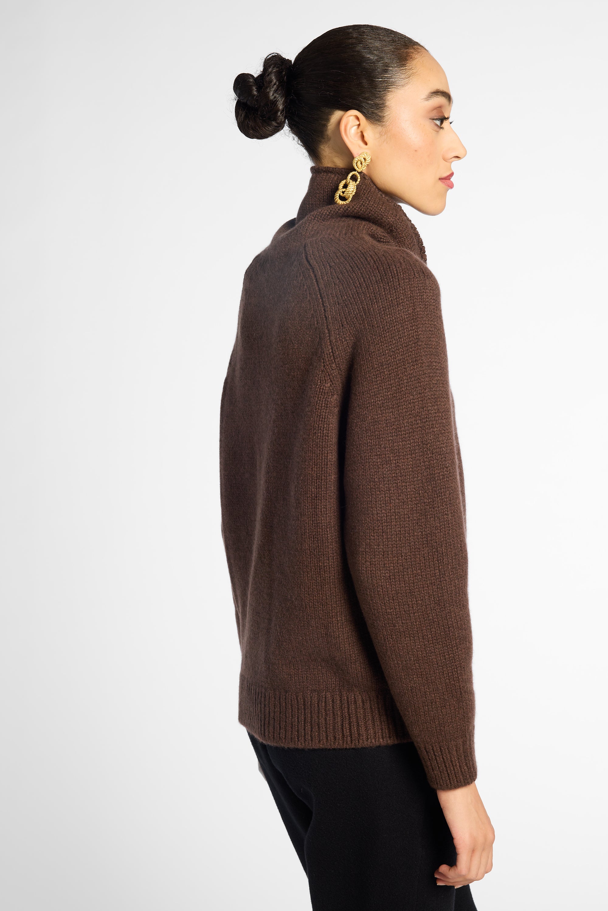 Side view: woman wearing a turtleneck sweater in Chocolate brown with black pants in front of a white background 