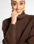 Close-up view: woman wearing a turtleneck sweater in Chocolate brown with black pants in front of a white background 