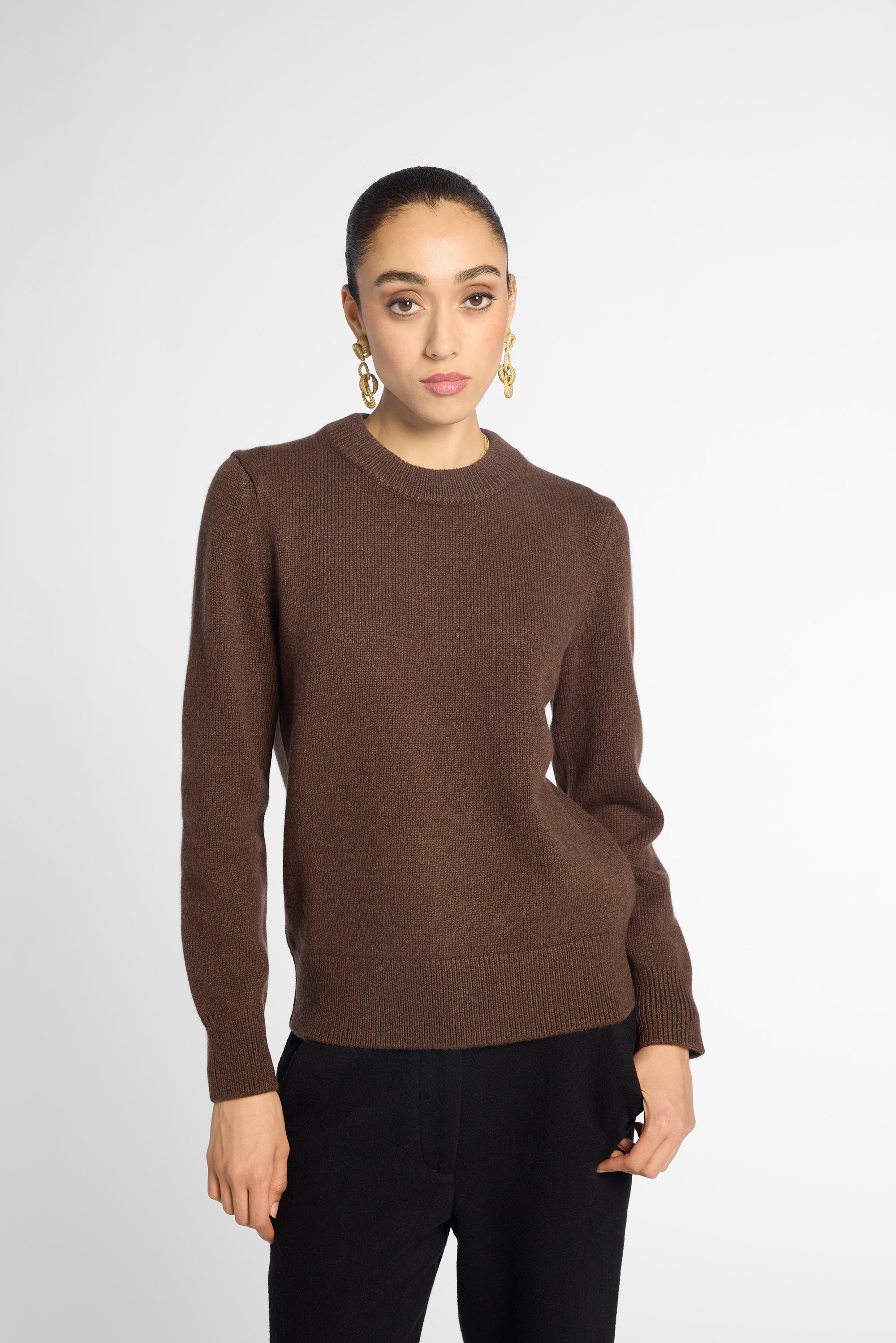 Front view: woman wearing a classic crewneck sweater in Chocolate brown with black pants in front of a white background 