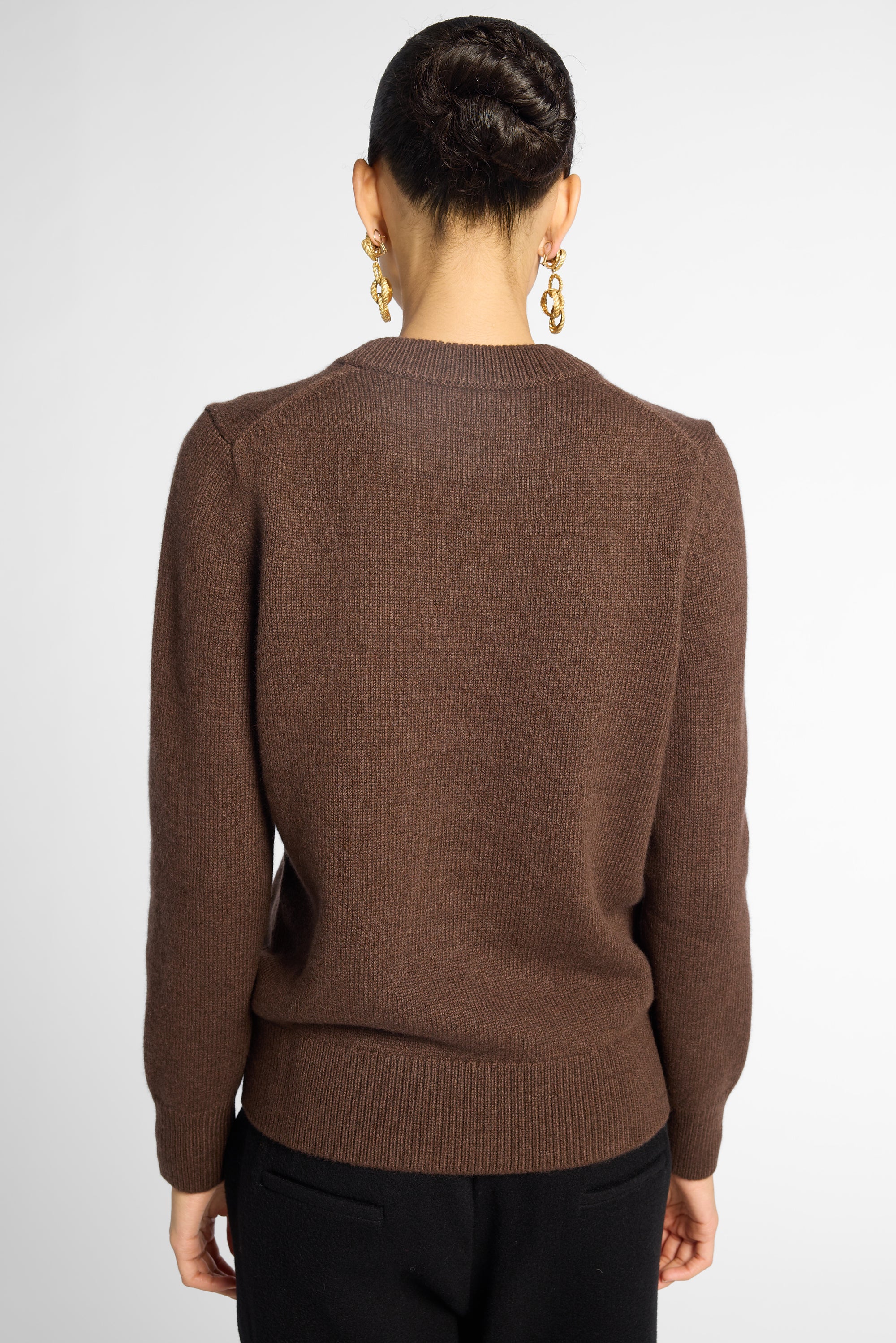 Back view: woman wearing a classic crewneck sweater in Chocolate brown with black pants in front of a white background 