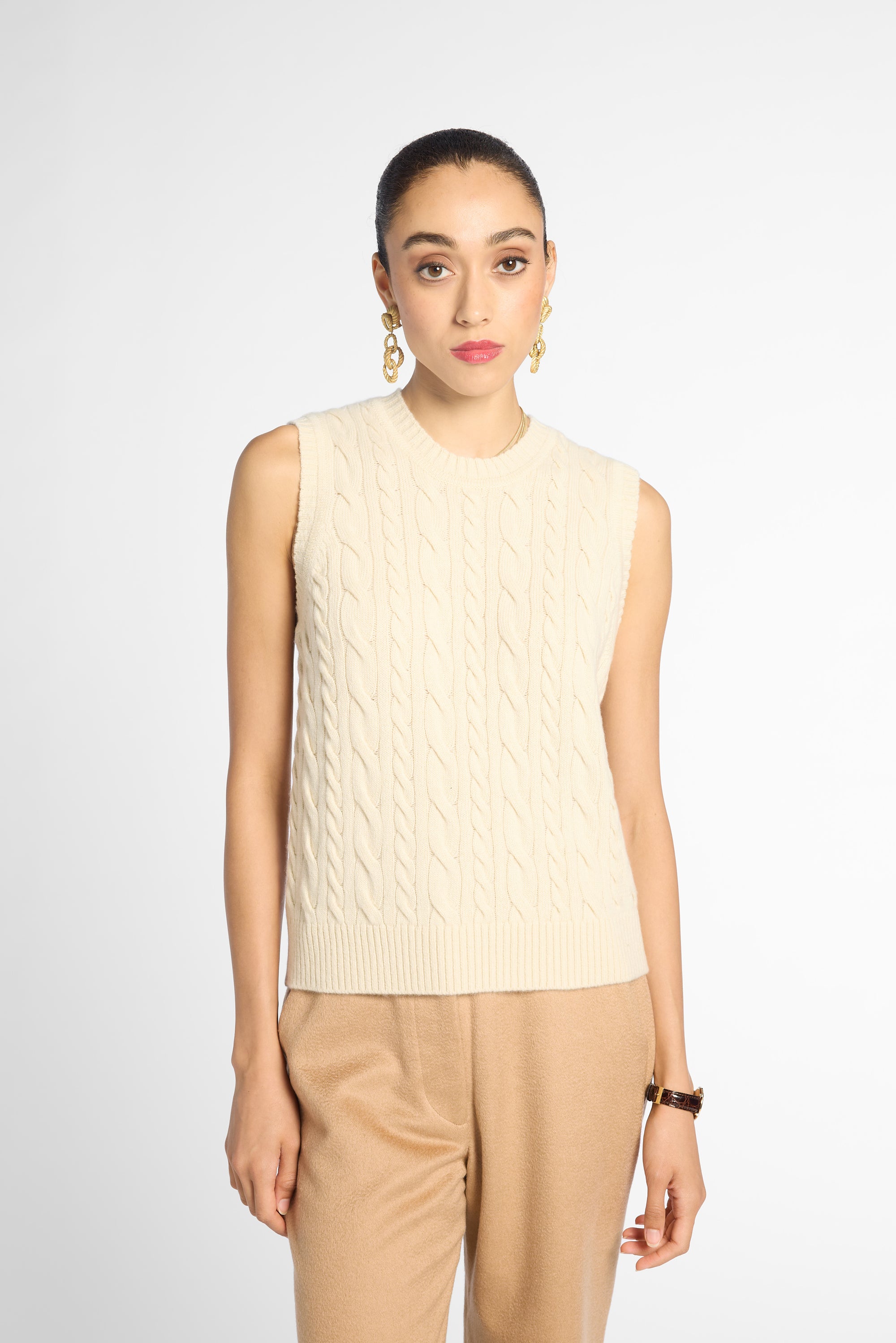 A woman modeling a sleeveless cream cable-knit sweater vest paired with tan trousers, photographed against a clean light gray studio background.