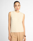 A woman modeling a sleeveless cream cable-knit sweater vest paired with tan trousers, photographed against a clean light gray studio background.