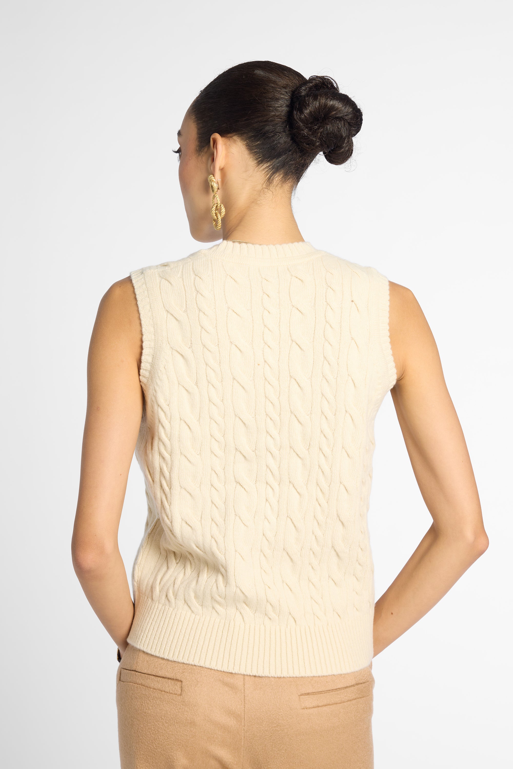 A woman shown from the back wearing a cream cable-knit sleeveless sweater vest and tan pants against a light gray studio background.