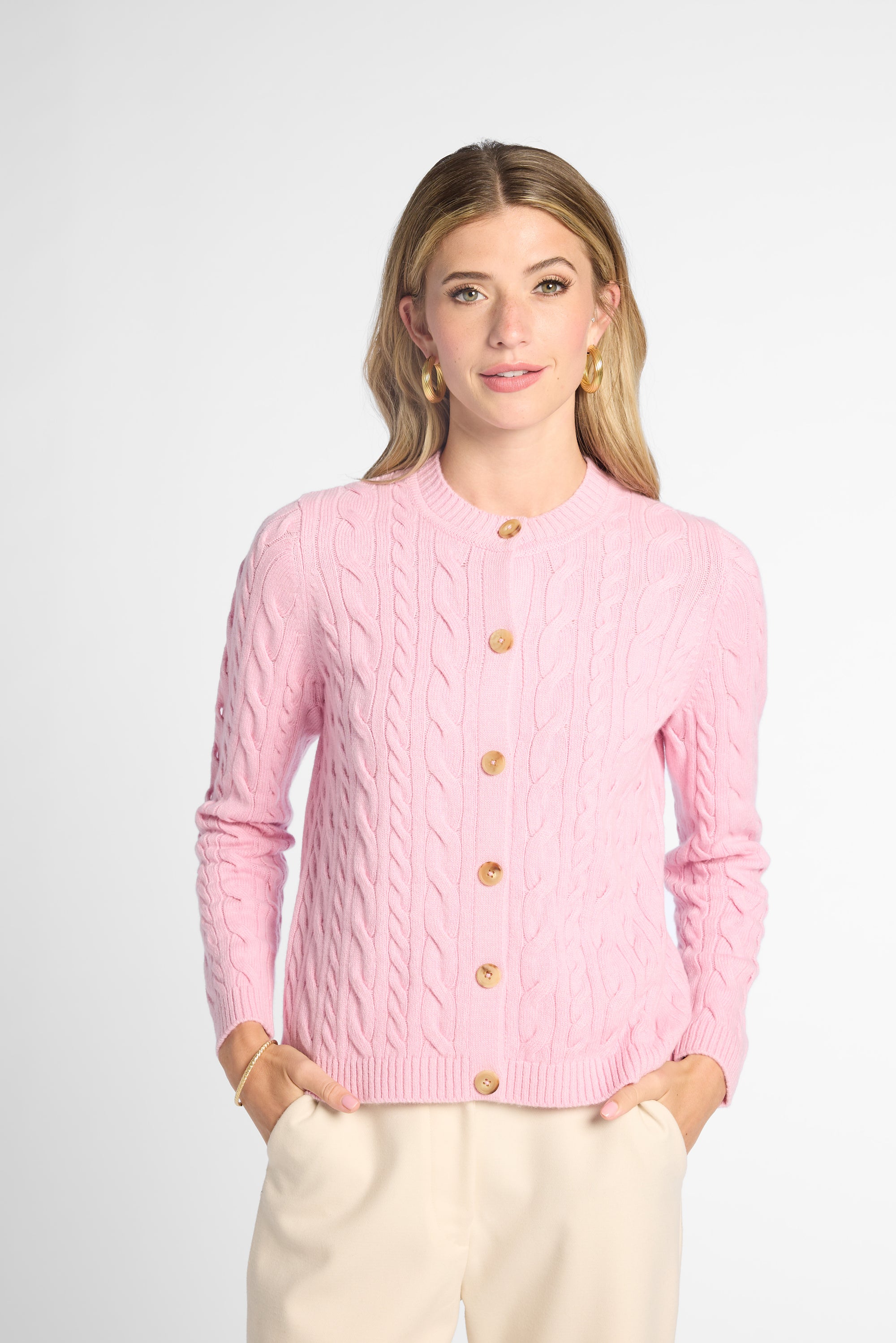A woman modeling a light pink cable-knit cardigan sweater with gold buttons, paired with cream trousers, against a light gray studio background.