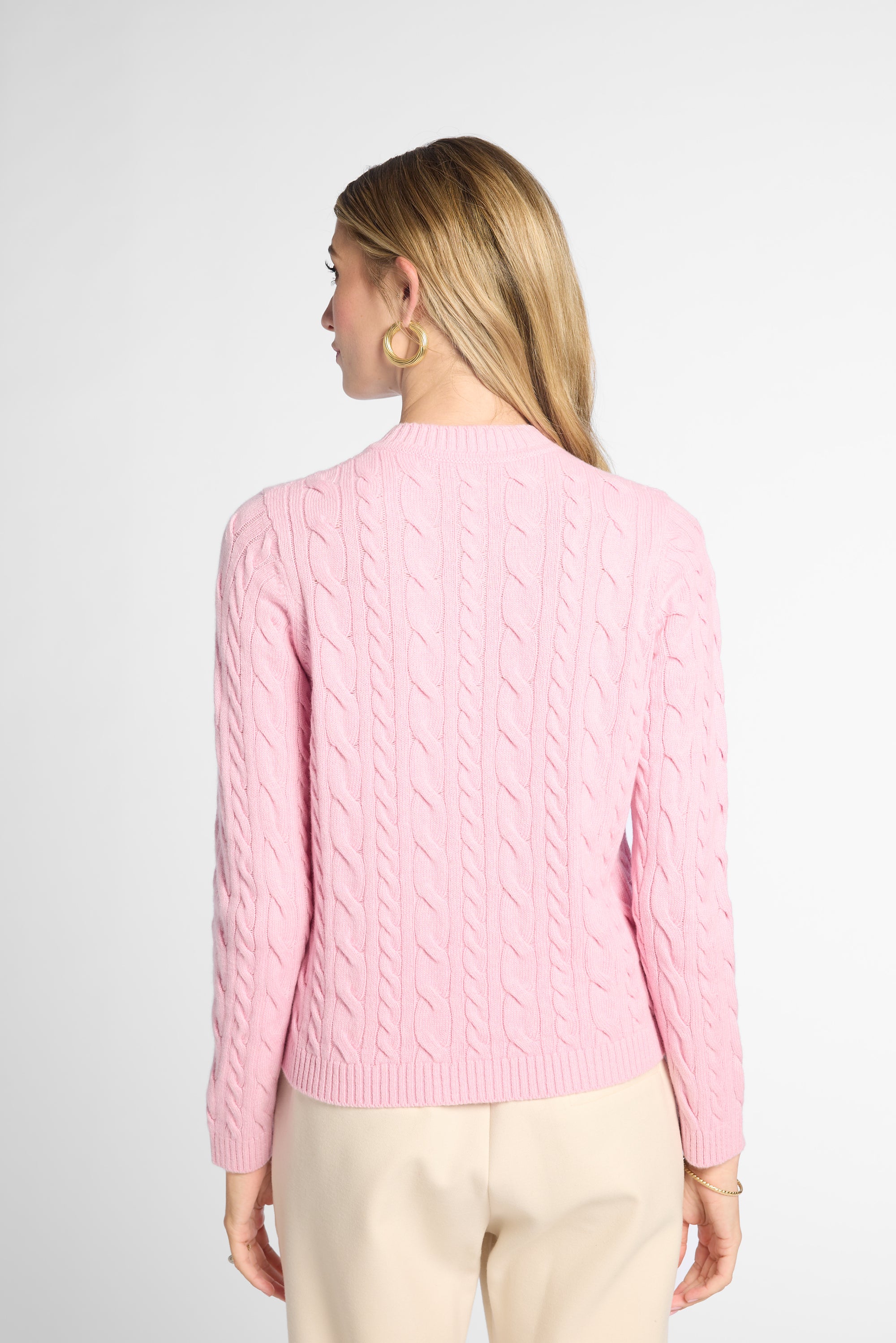 A woman shown from the back wearing a light pink cable-knit sweater and cream-colored trousers, photographed against a clean, light gray studio background.