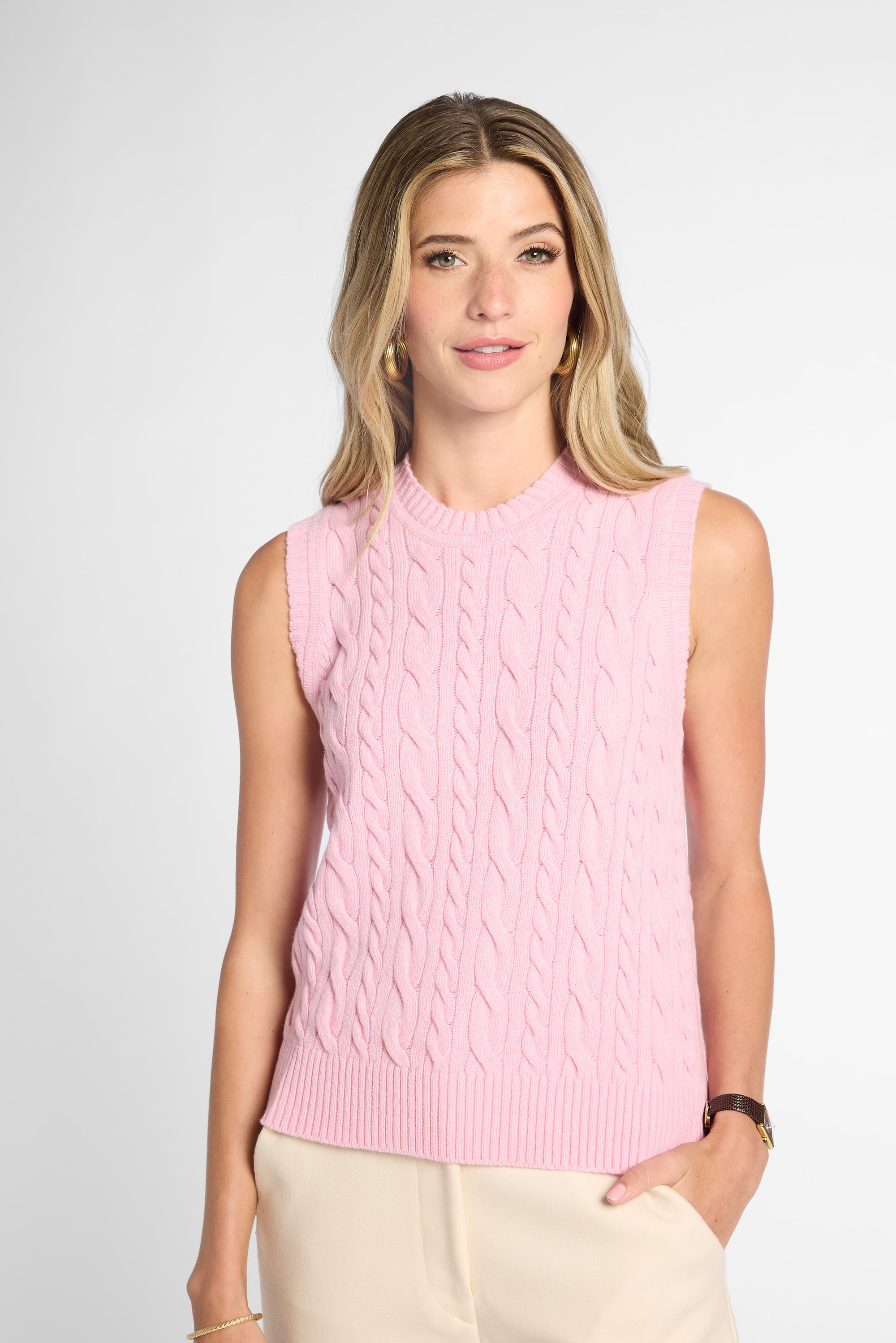 A woman modeling a sleeveless light pink cable-knit sweater vest against a light gray studio background.