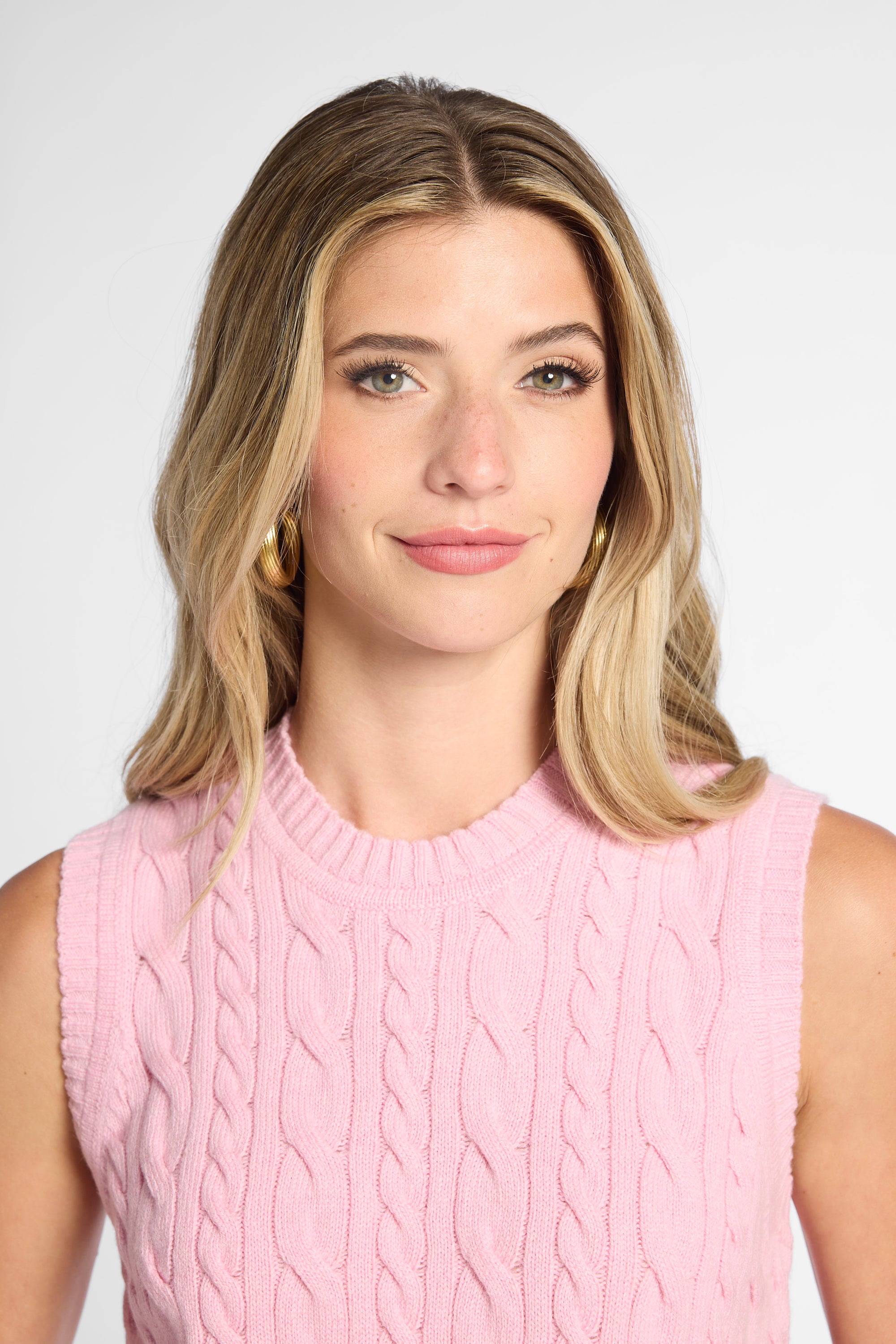 A woman with blonde hair wearing a pink sleeveless cable-knit sweater vest, photographed against a light gray studio background.