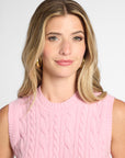 A woman with blonde hair wearing a pink sleeveless cable-knit sweater vest, photographed against a light gray studio background.