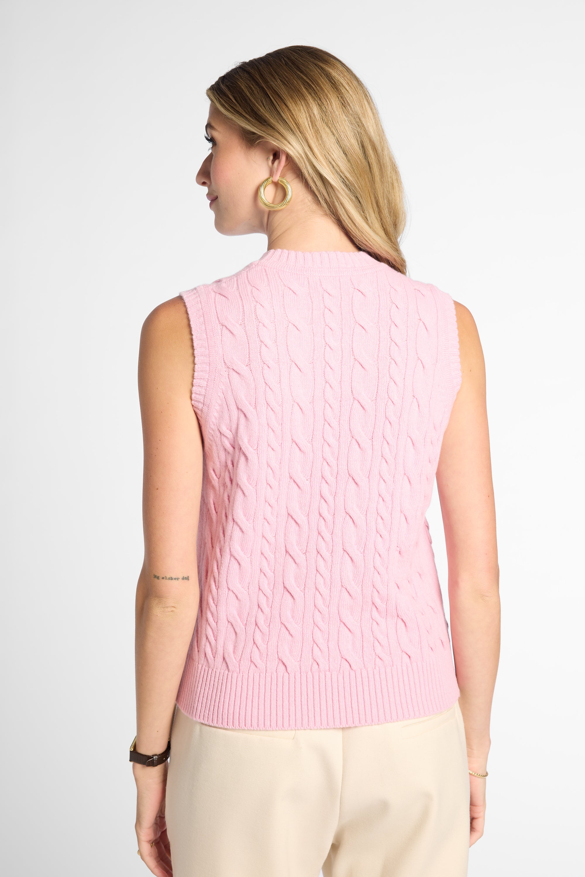 A woman shown from the back wearing a sleeveless pink cable-knit sweater vest and light beige pants against a plain light gray studio background.