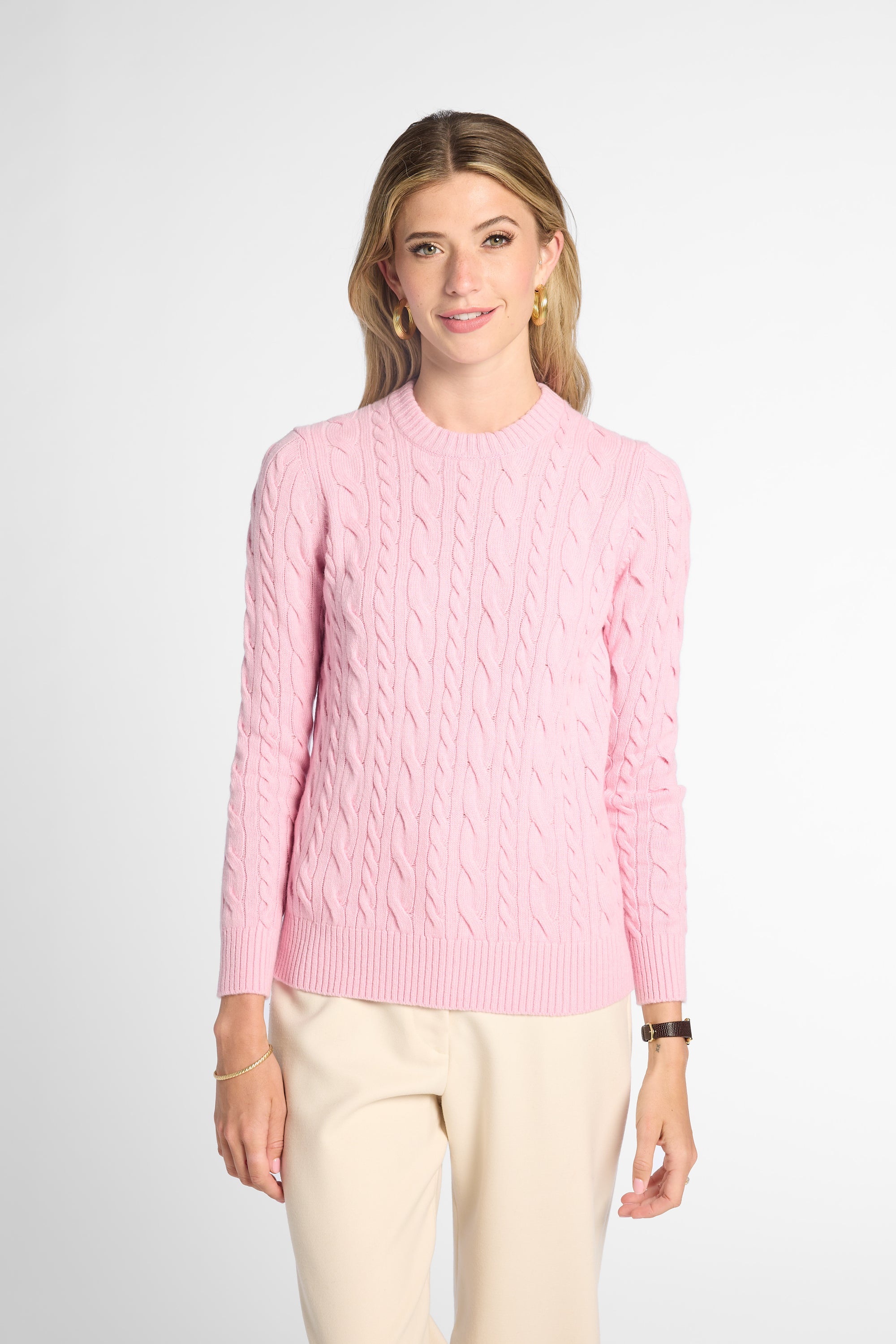 A woman wearing a light pink cable-knit crewneck sweater and cream-colored trousers, standing against a plain light gray studio background.