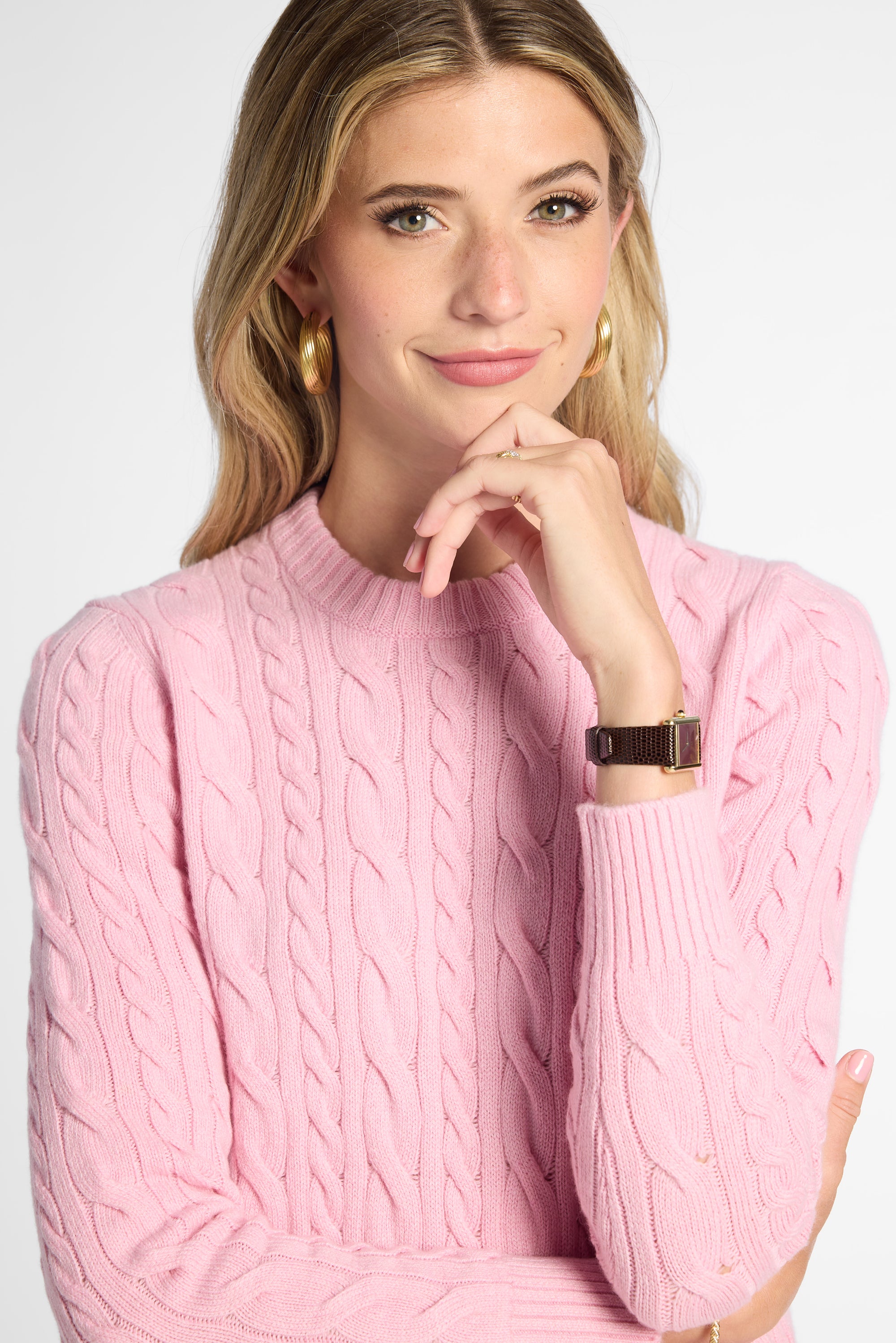 A woman wearing a pink cable-knit mock neck sweater, posing with her hand near her chin against a light gray background.